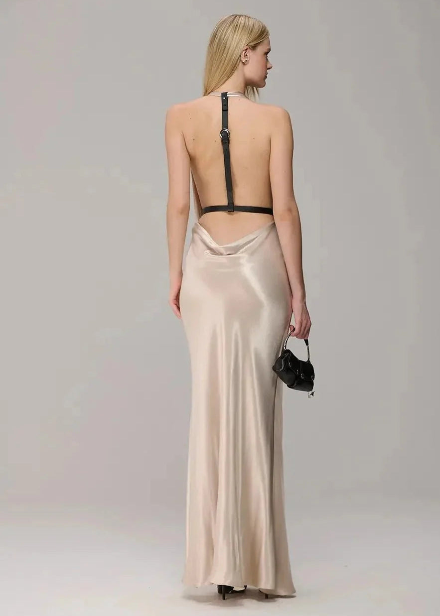 A woman shown from the back wearing a floor-length satin slip dress in a warm champagne beige color with a black strap detail at the open back. She is holding a small black handbag against a plain light gray studio background.