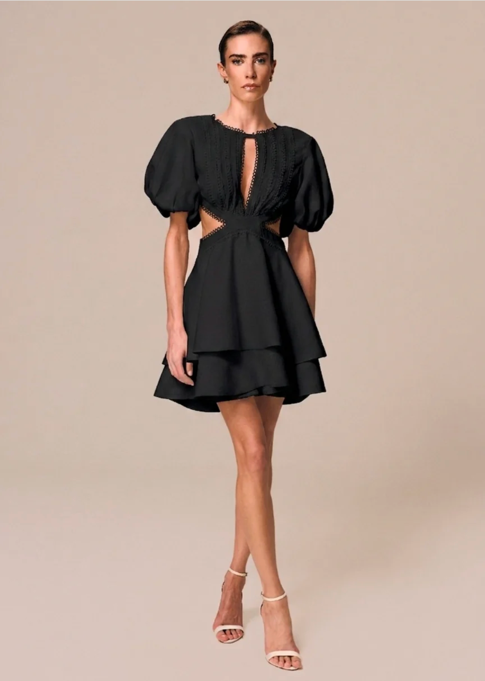 A model wearing a black puff-sleeve mini dress with a cutout waist and lace-up keyhole neckline, paired with beige strappy high-heeled sandals, set against a solid beige studio background.