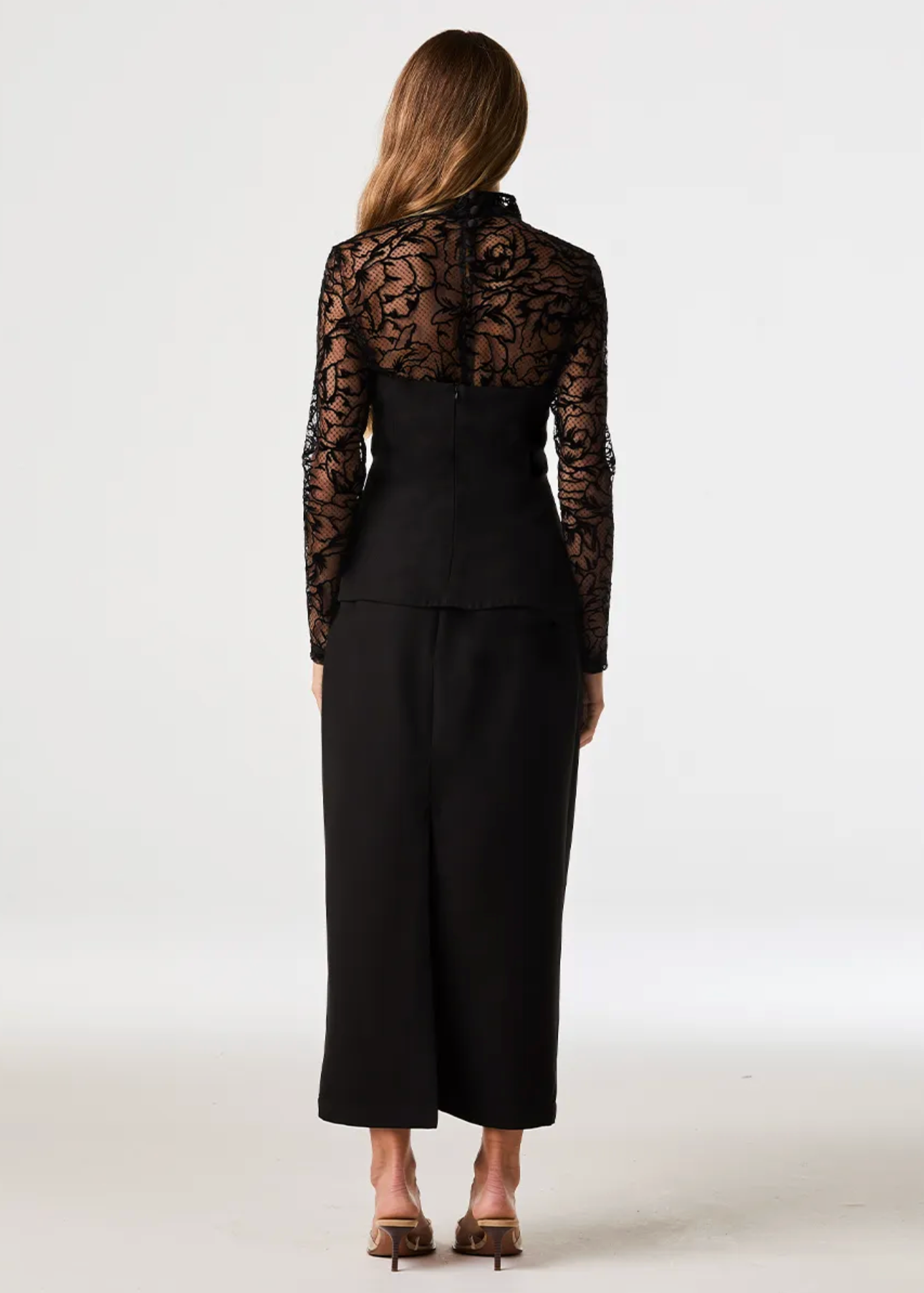 A woman viewed from the back wearing a black long-sleeve lace top with a structured waist and wide-leg black pants, standing against a plain light studio background.