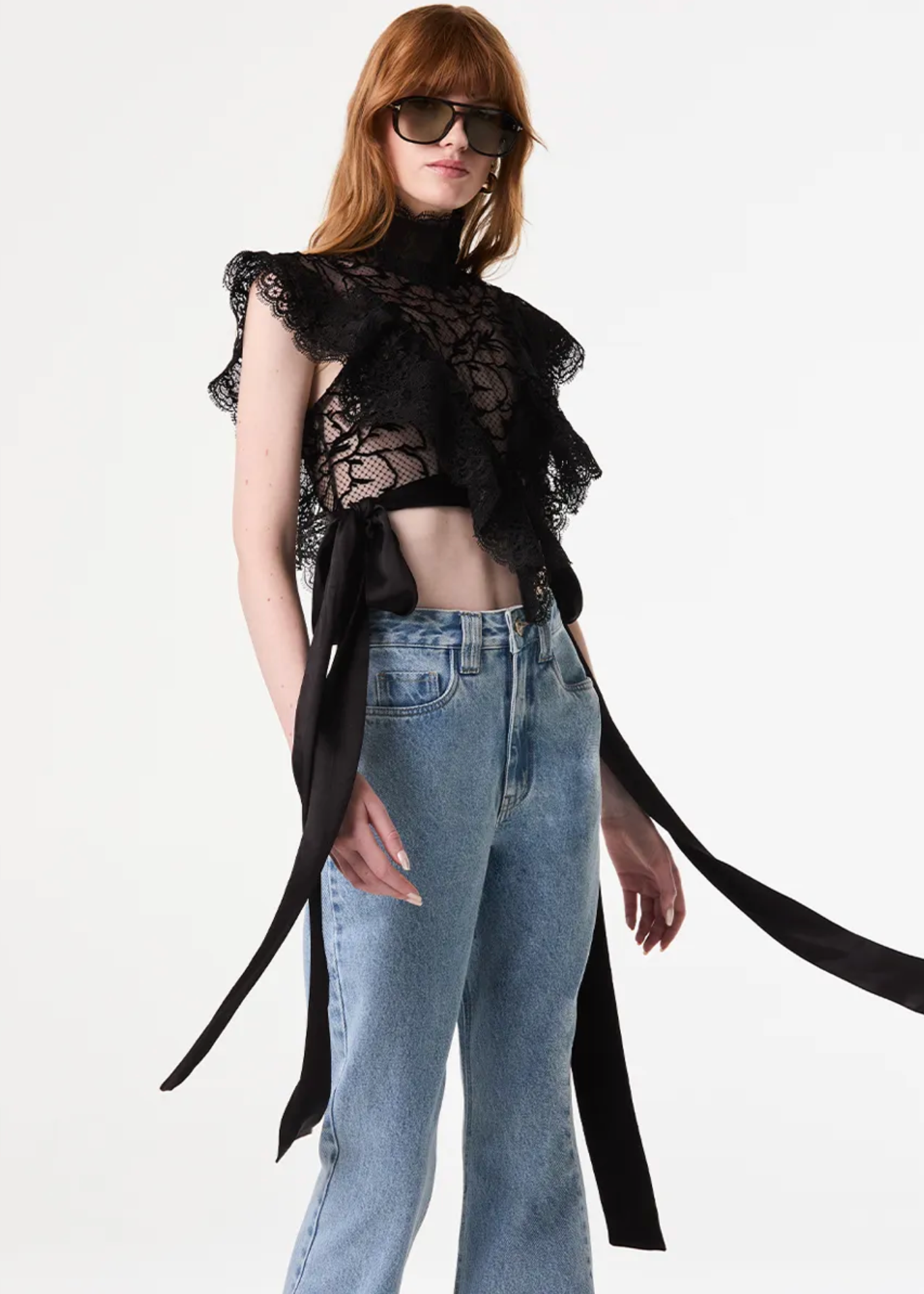 A model wearing a black lace ruffled crop top with long ribbon ties and light-wash blue jeans, standing against a plain white studio background.