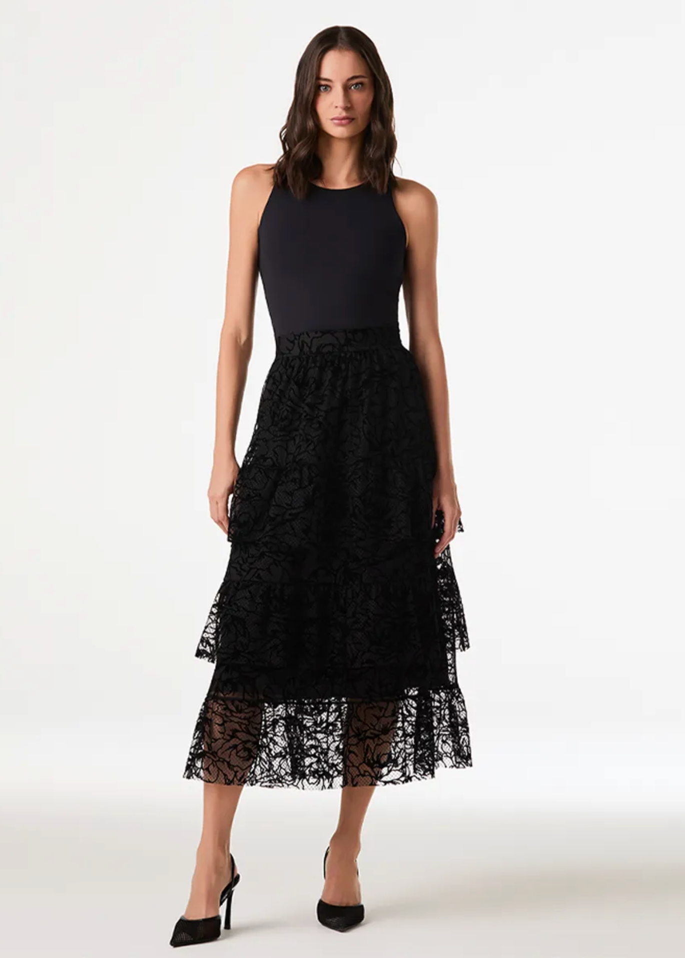 A model wearing a sleeveless black midi dress with a solid black bodice and a layered black lace tiered skirt, paired with black high heels, against a plain white studio background.
