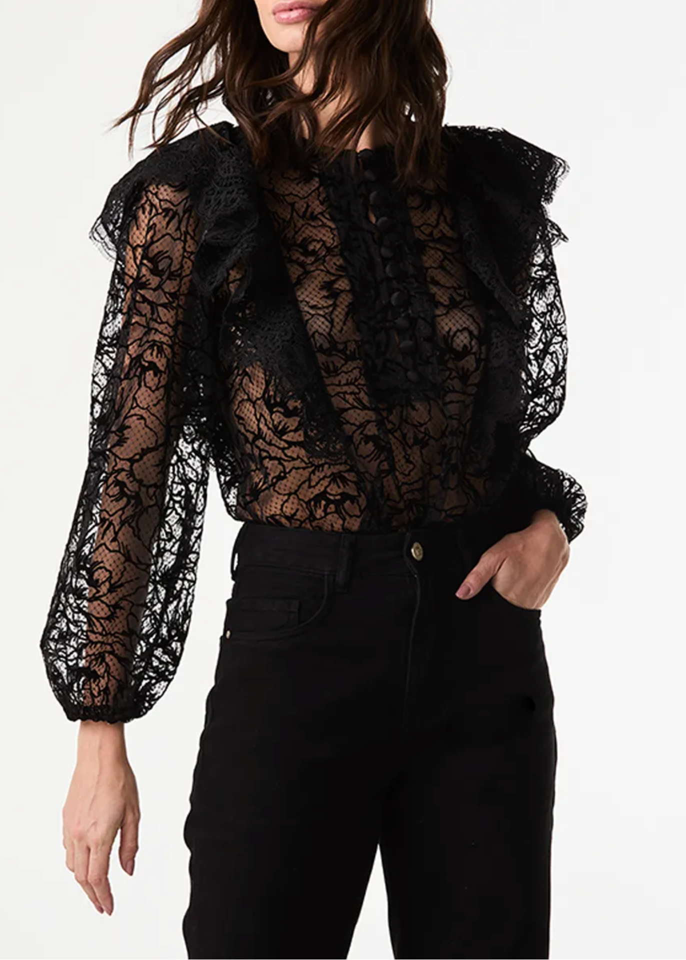 A woman wearing a black sheer lace blouse with dramatic ruffled shoulders and long balloon sleeves, paired with black jeans, against a plain white studio background.