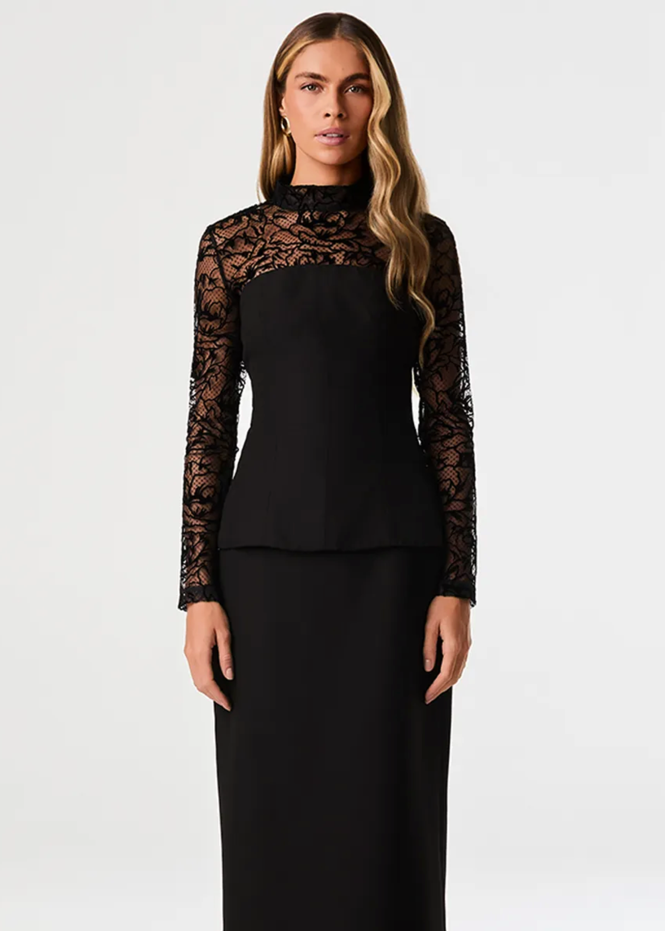 A woman modeling a black long-sleeve dress with sheer lace sleeves and a lace yoke, paired with a matching black skirt, against a plain light gray studio background.