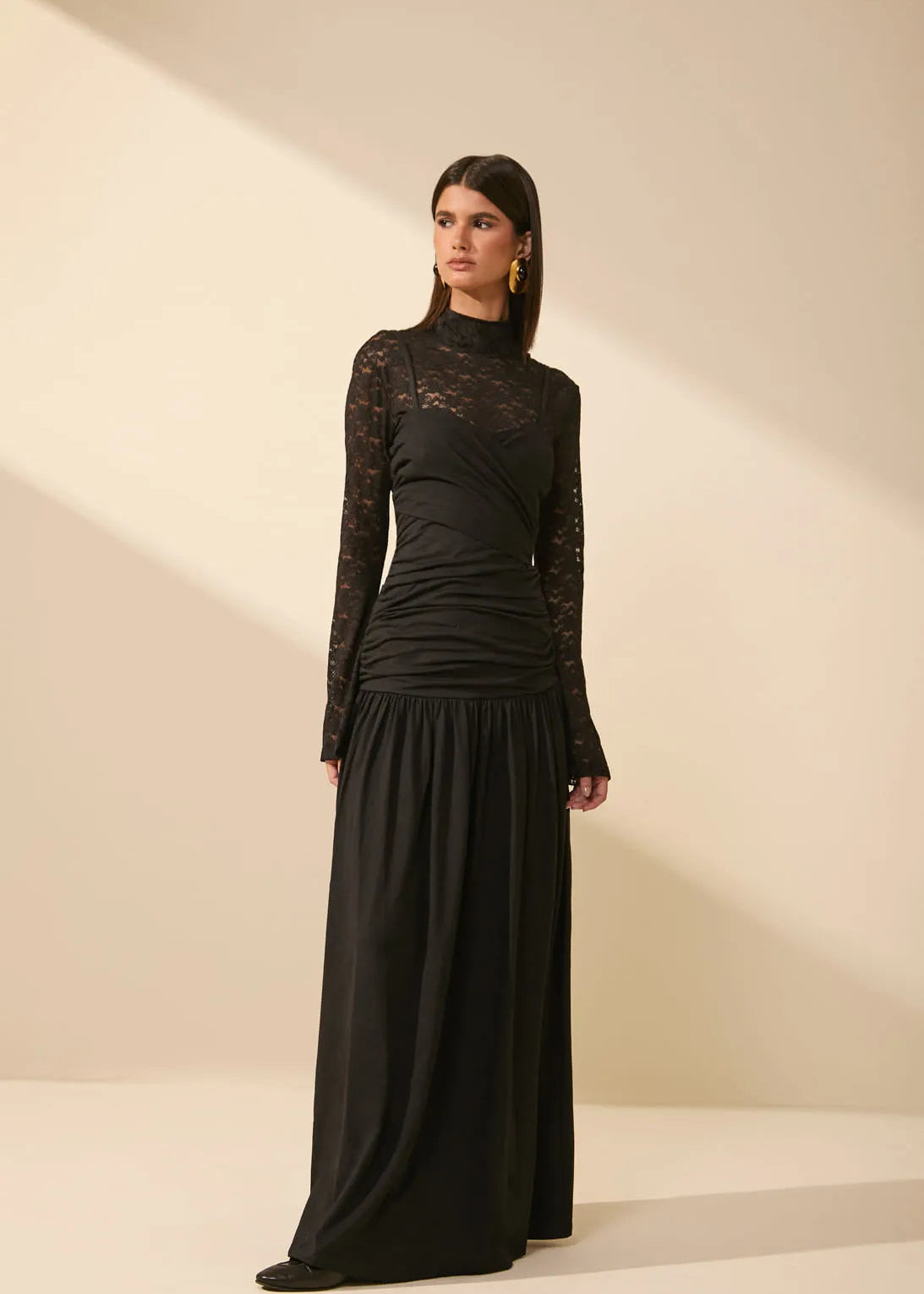 A woman modeling a long black dress with sheer lace sleeves and a gathered ruched bodice, standing against a warm beige studio backdrop with soft diagonal shadows.