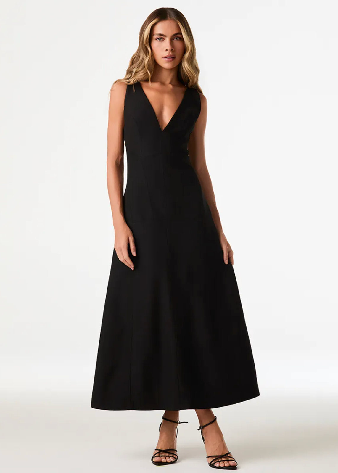 A woman modeling a sleeveless black midi dress with a deep V-neckline and flared skirt, paired with black strappy heels, against a plain white studio background.