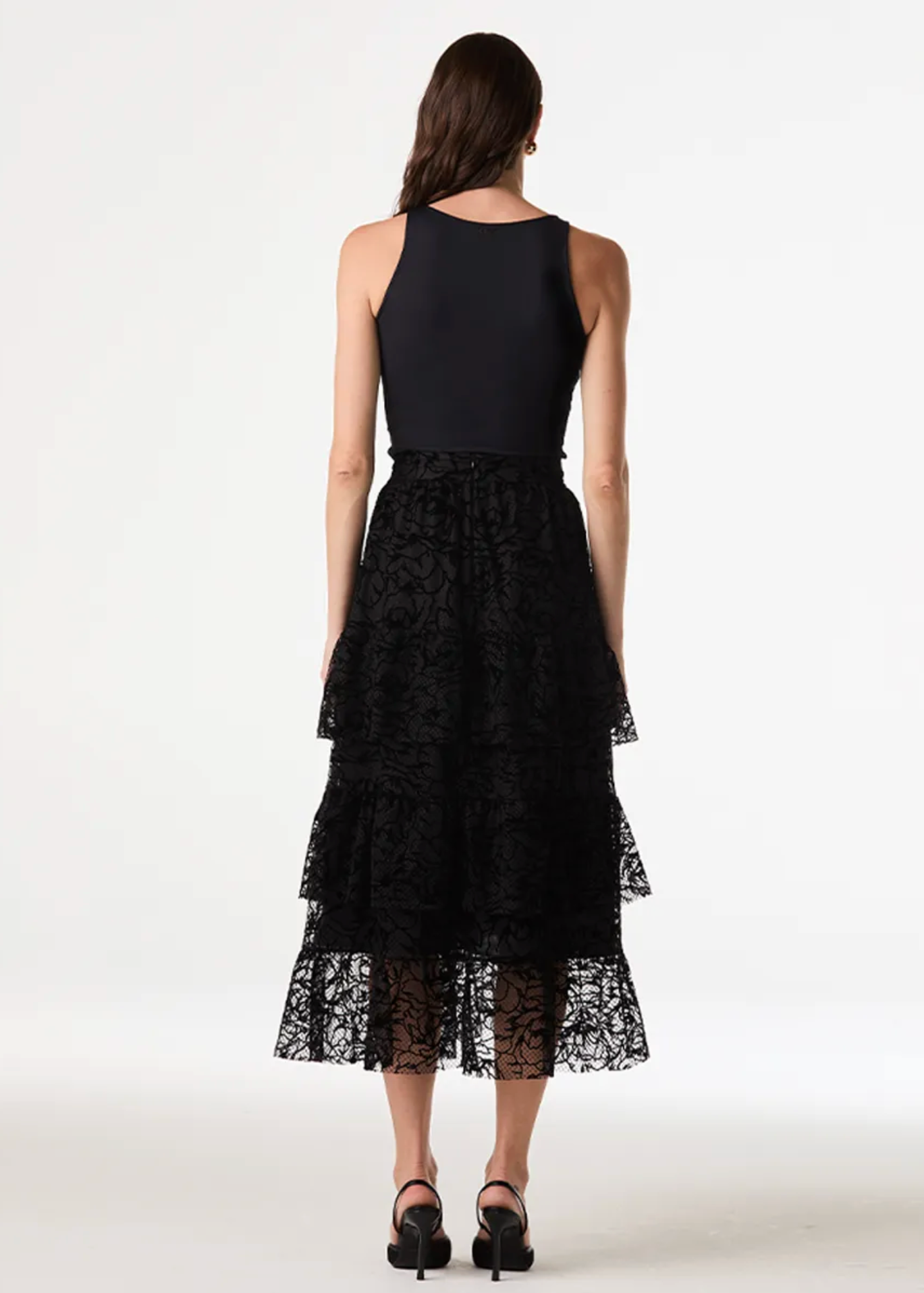 A woman viewed from the back wearing a sleeveless black dress with a solid black bodice and a layered black lace skirt, photographed against a clean white studio background.