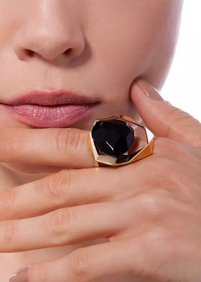 Close-up of a hand wearing a gold ring with a black gemstone, touching the face.