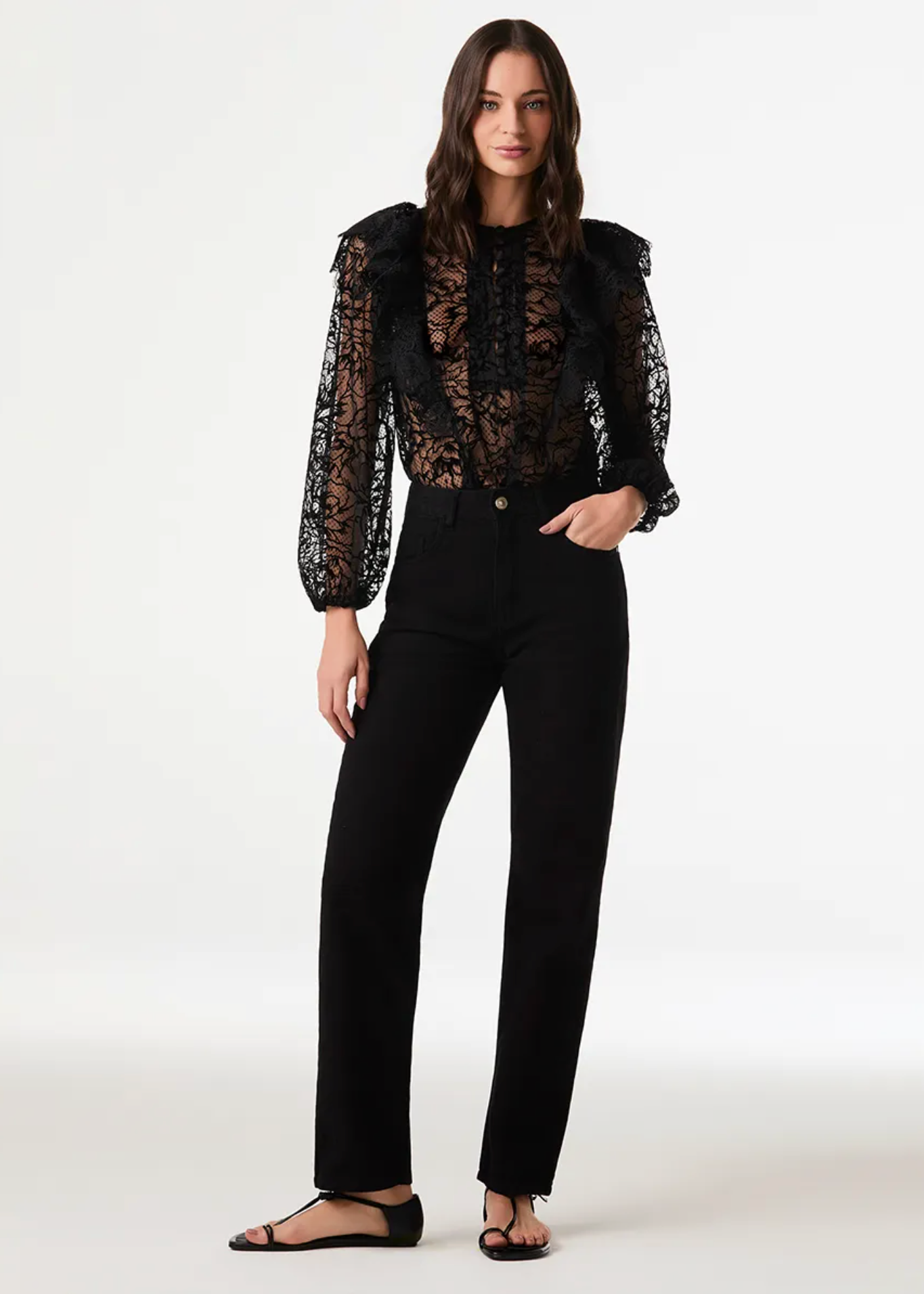 A model stands against a plain light gray studio background wearing a black lace long-sleeve top with ruffled shoulders and black straight-leg pants.