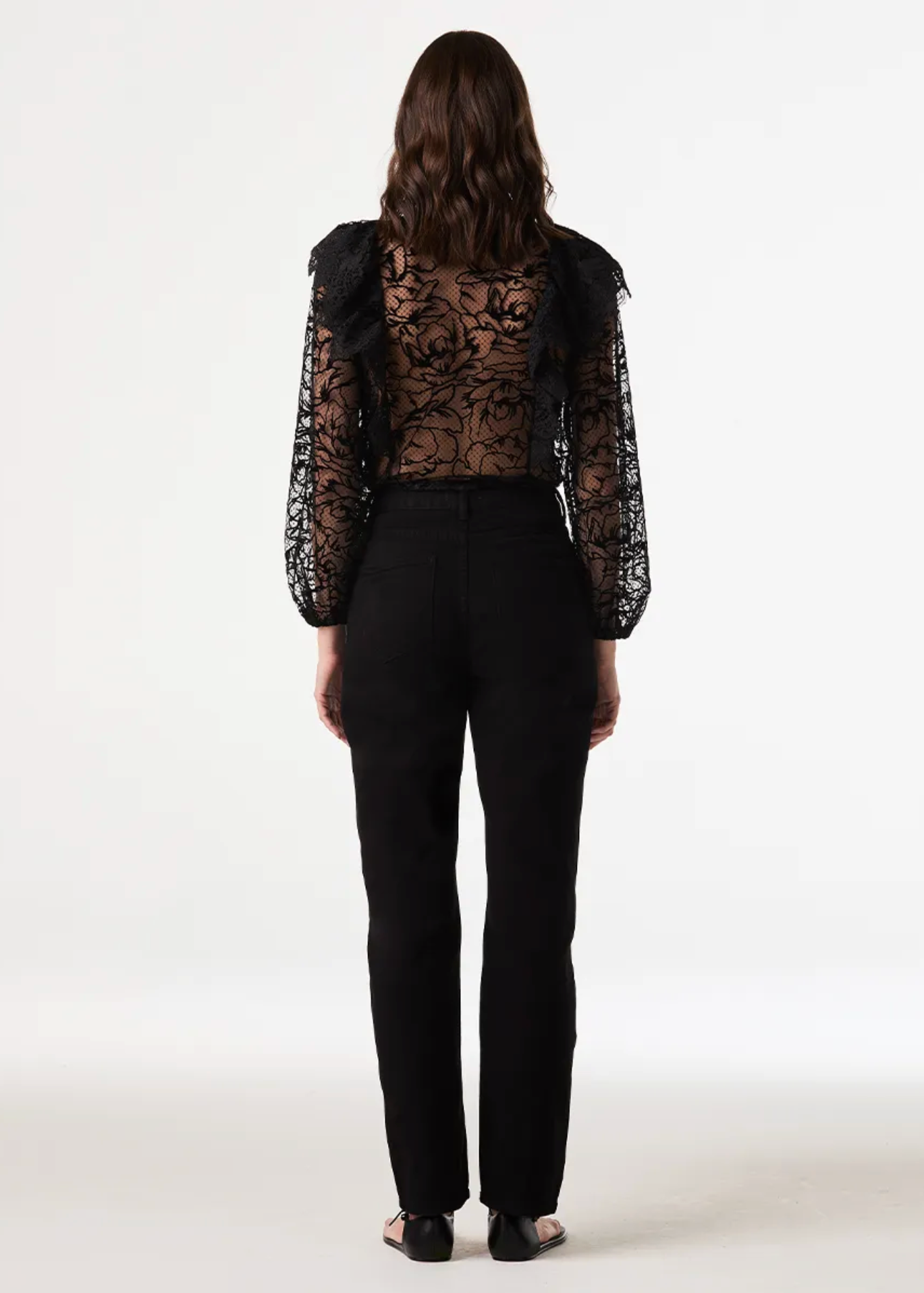 A woman viewed from the back wearing a black sheer floral lace long-sleeve top with ruffled shoulders and black pants, standing against a plain white studio background.