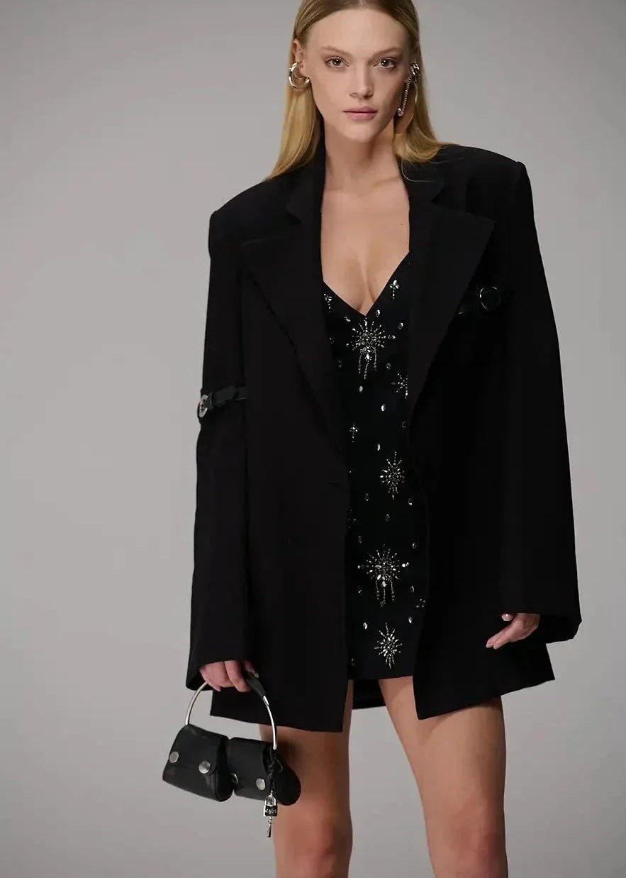 Woman wearing a black outfit with a starry pattern, holding a small black handbag against a gray background.