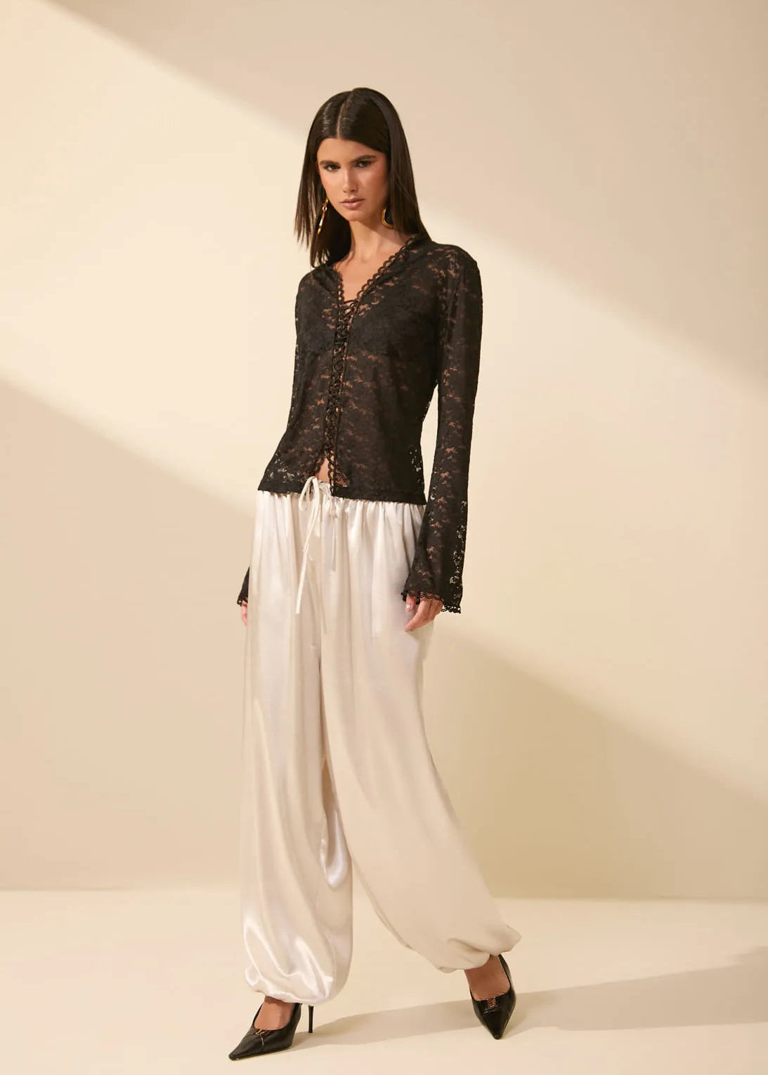 A model wearing a black lace long-sleeve top and shiny cream satin wide-leg pants, standing against a warm beige studio background with diagonal shadows.