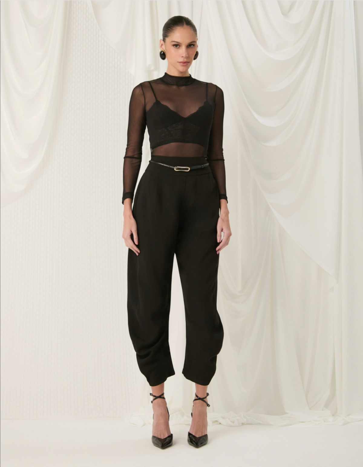 A model stands against a draped white fabric backdrop wearing a black sheer long-sleeve mock-neck top with a black lace insert, paired with black high-waisted balloon pants and black strappy heels.