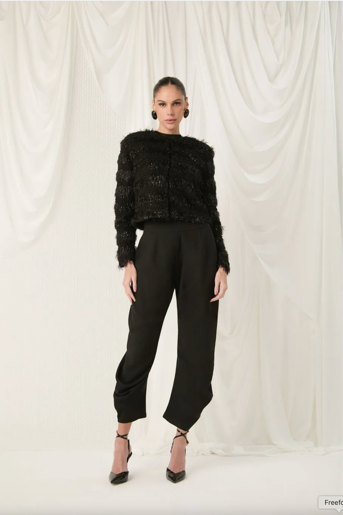 A model stands against a draped white fabric backdrop wearing a black textured fuzzy cropped top, black wide-leg cropped pants, and black high heels.