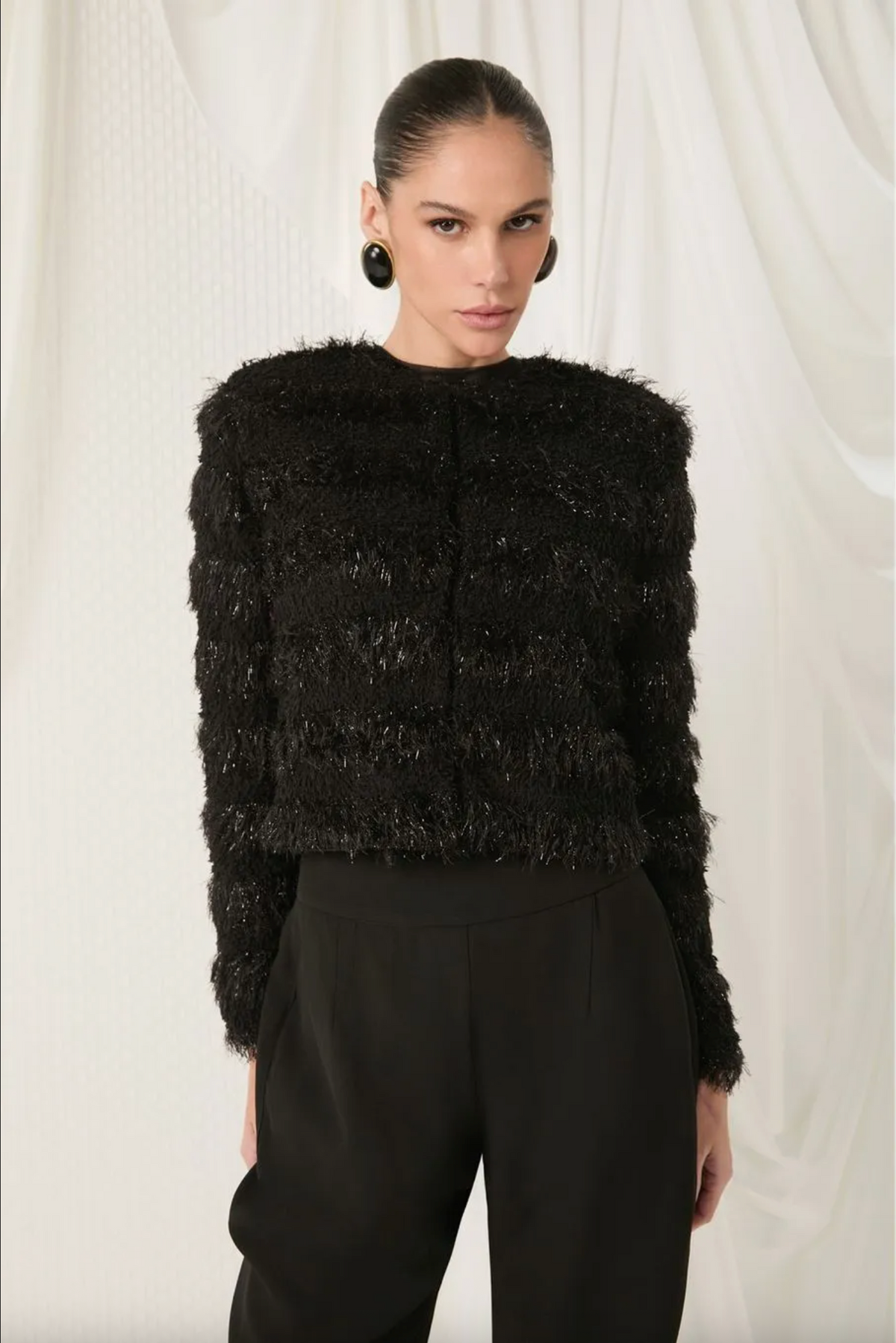 A model wearing a black textured, fuzzy cropped jacket with long sleeves and black trousers, standing against a draped white fabric background.