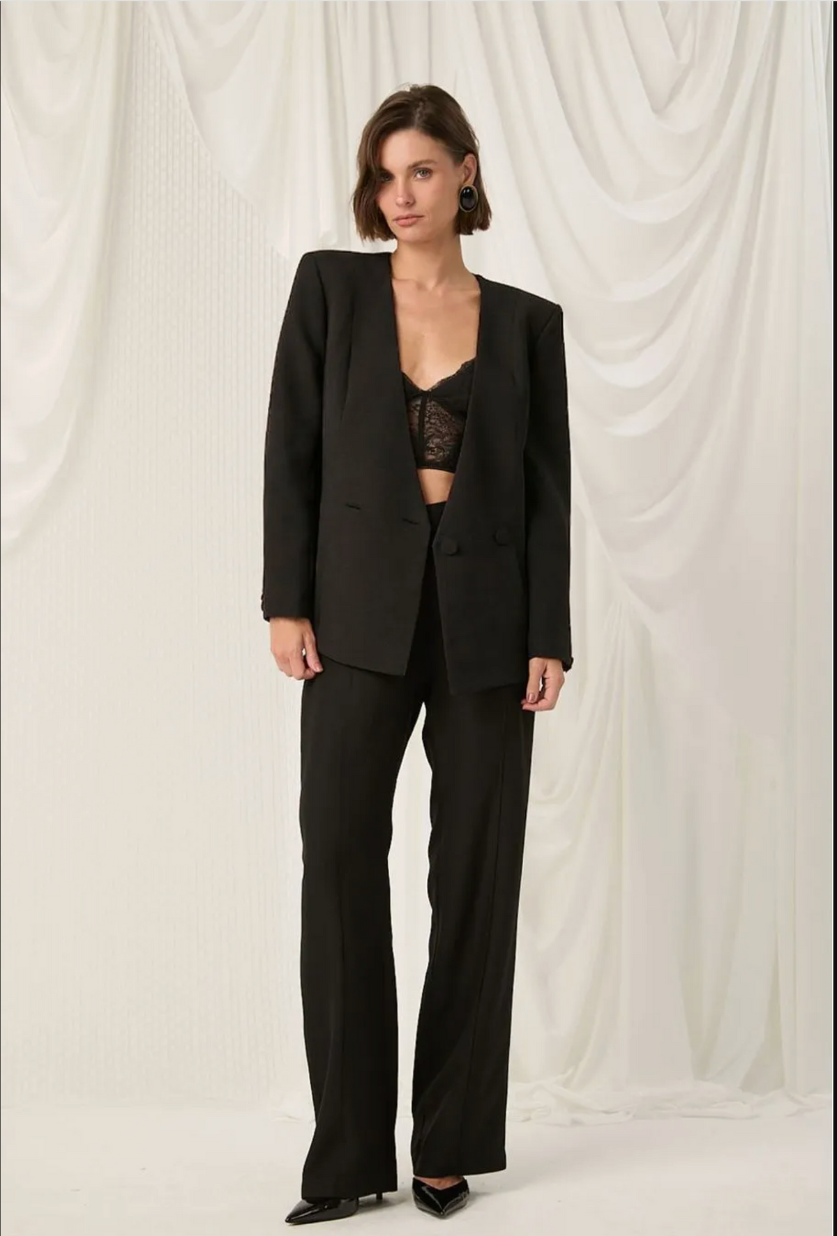 A model stands against a draped white fabric backdrop wearing a black tailored blazer and matching wide-leg trousers over a black lace crop top.