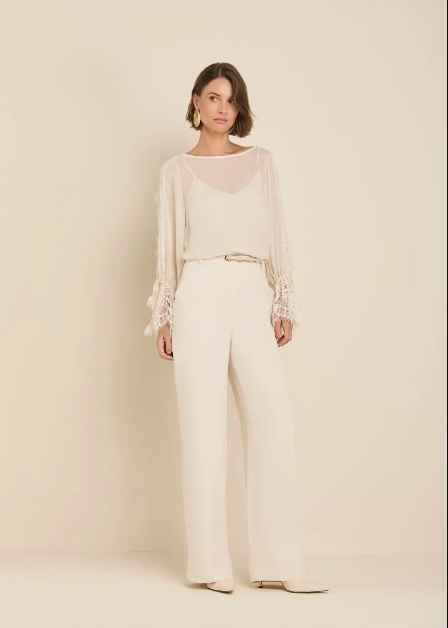 A woman stands against a plain warm beige studio background wearing a coordinated cream outfit: a sheer long-sleeve top with lace trim at the cuffs, a matching camisole, a thin belt, and wide-leg trousers.