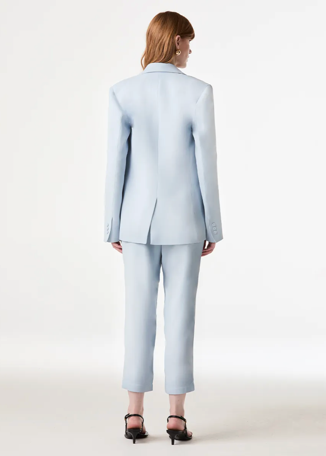 A model viewed from the back wearing a light blue tailored suit with a single-breasted blazer and cropped trousers, paired with black heeled sandals against a clean white studio background.