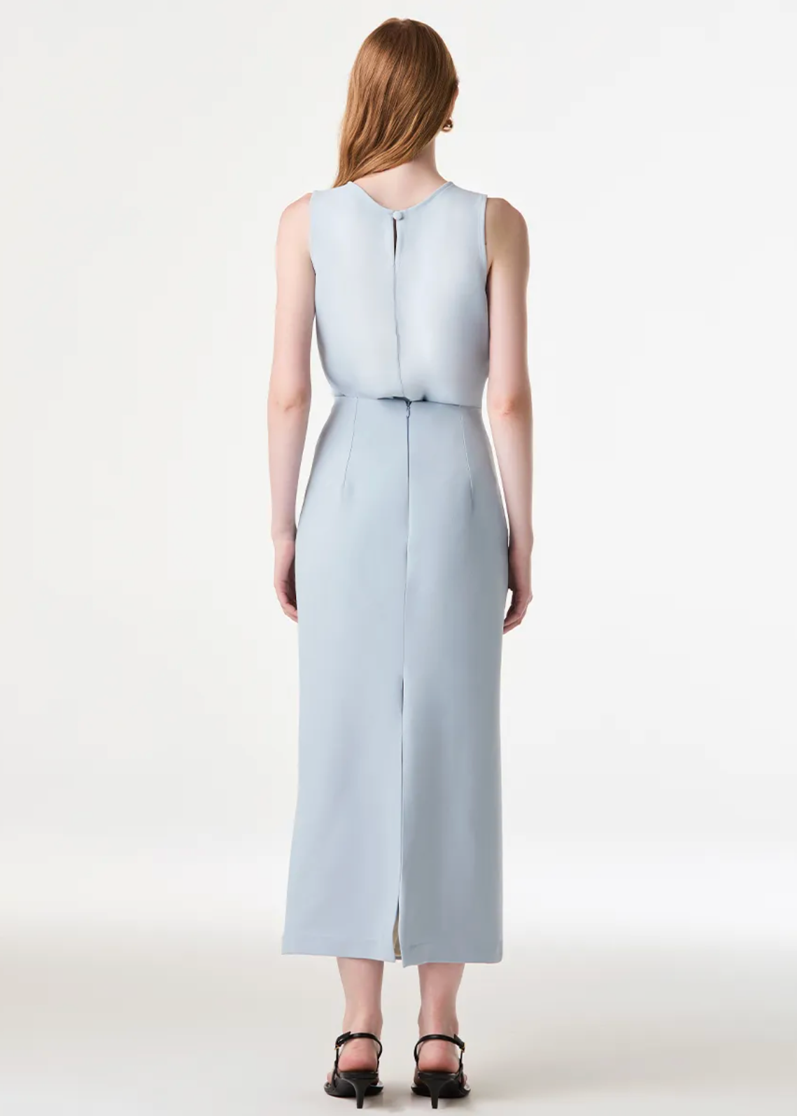 A woman viewed from the back wearing a sleeveless light blue midi dress with a tie waist and front slit, paired with black sandals, against a plain white studio background.