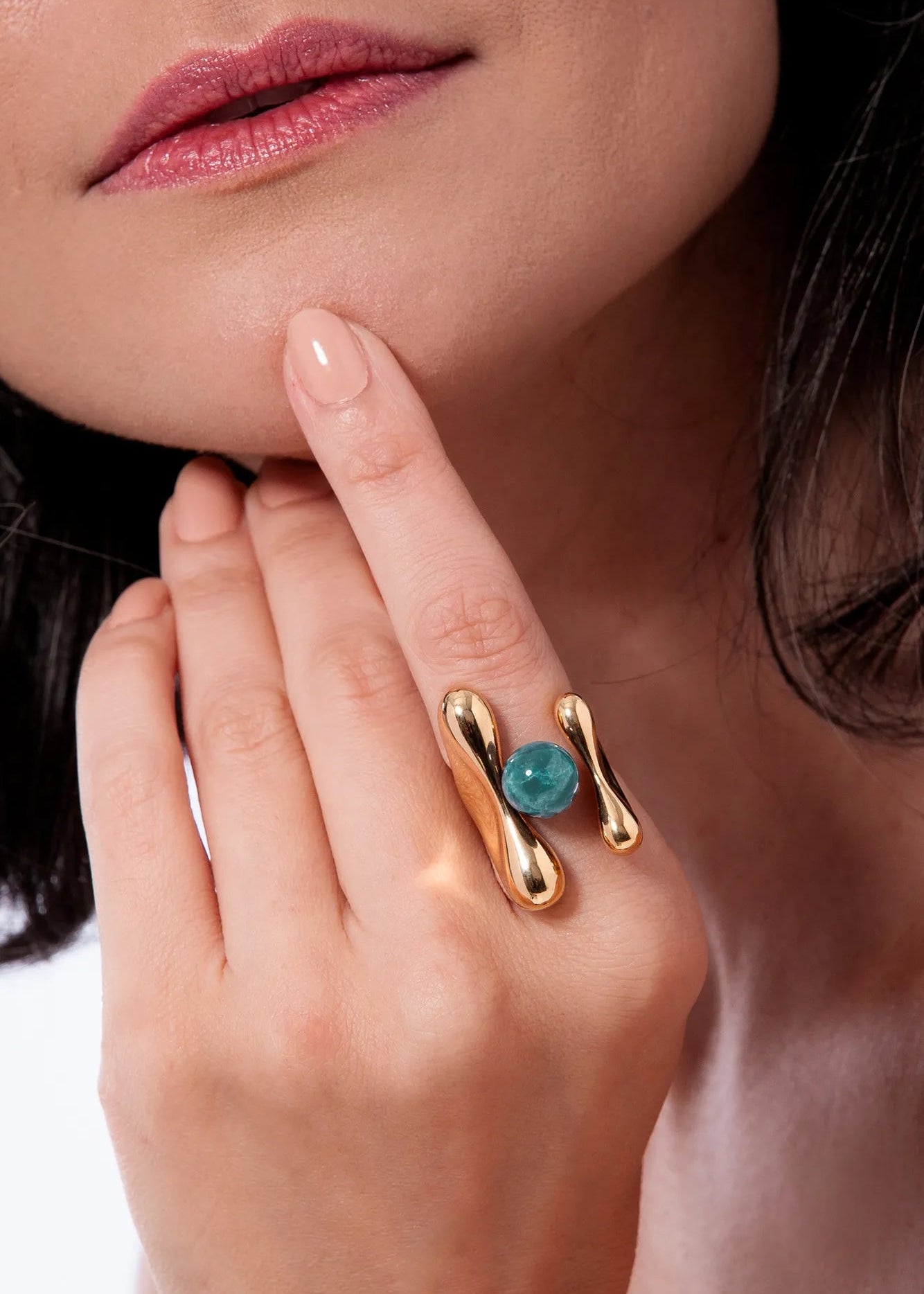 Close-up of a hand wearing a gold ring with a blue gemstone, touching the chin.