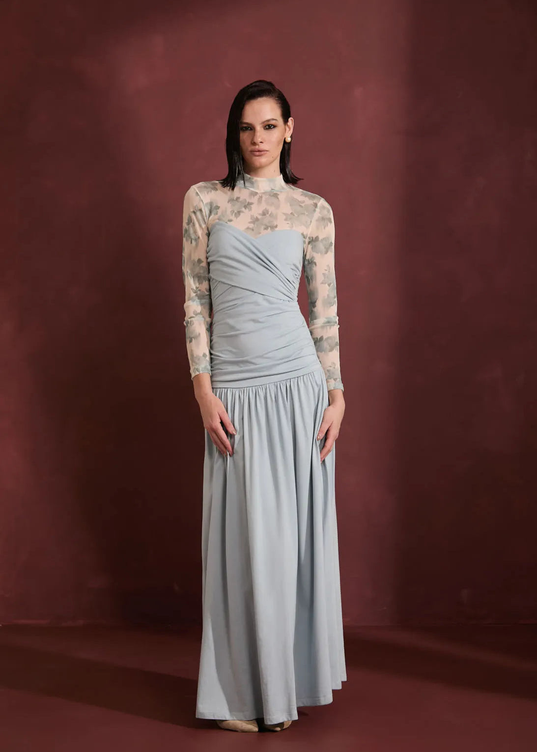 A woman models a floor-length formal gown with a light blue-gray draped skirt and sheer floral-patterned long sleeves, standing against a dark reddish-brown studio backdrop.