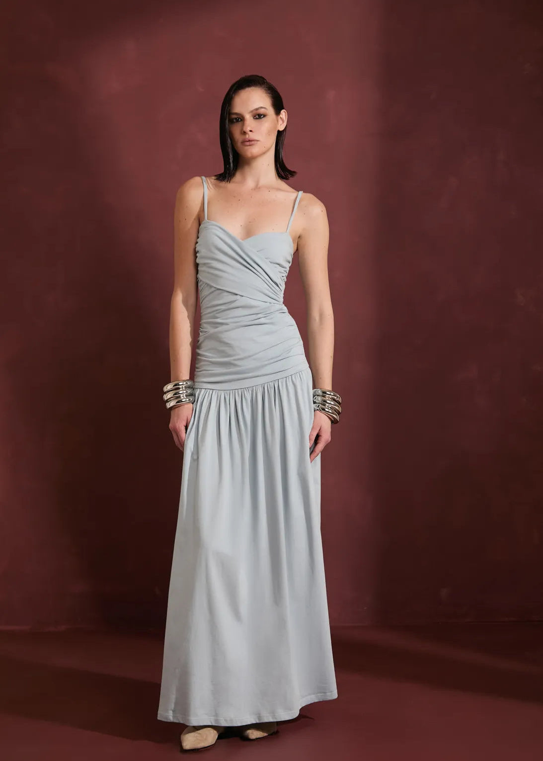 A woman modeling a floor-length light gray sleeveless gown with thin straps, a draped sweetheart neckline, and a gathered waist, standing against a dark reddish-brown studio backdrop.