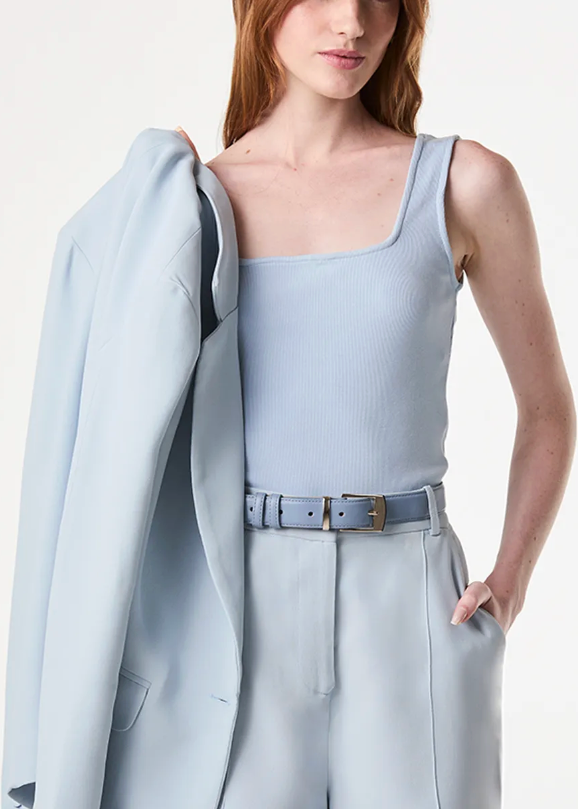 A model wearing a light blue ribbed tank top, matching wide-leg trousers, and a matching belt, with a light blue blazer draped over her shoulder against a plain white background.