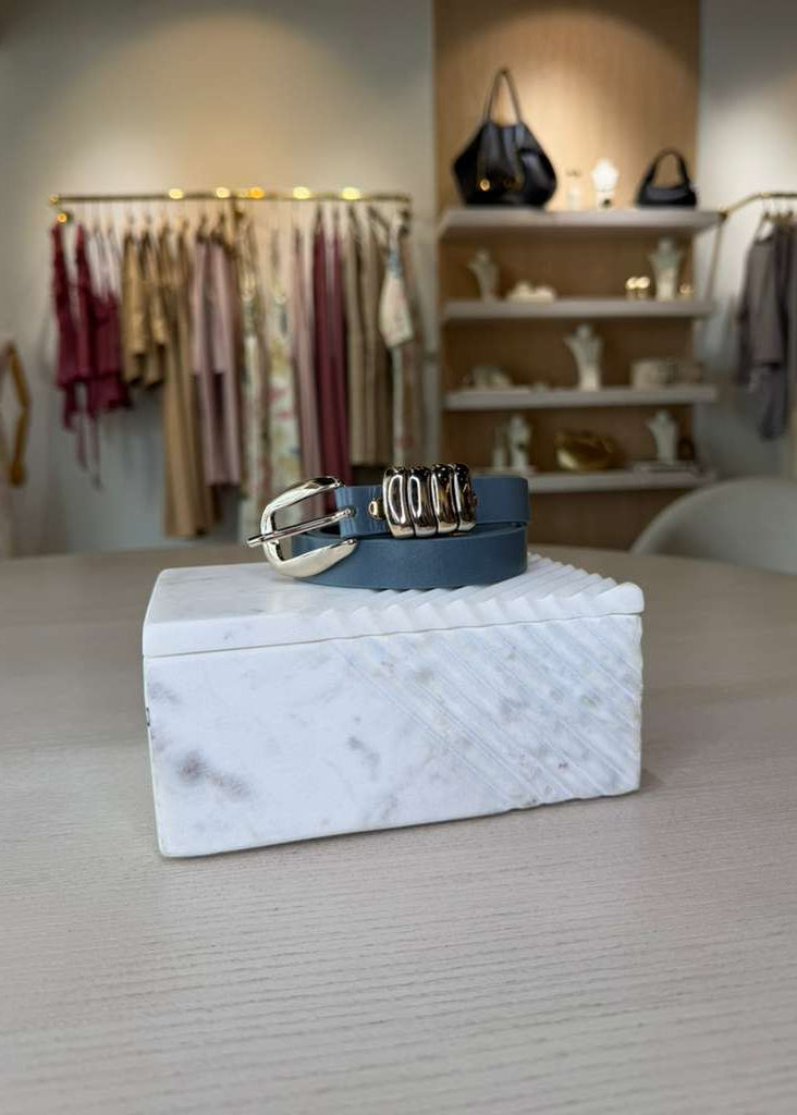 Blue leather belt with silver buckle on a white textured box in a clothing store setting.
