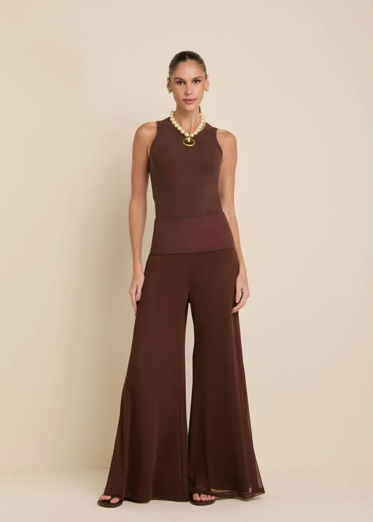 A model stands against a warm beige studio background wearing a sleeveless, high-neck, wide-leg jumpsuit in a rich dark brown color, styled with a chunky gold necklace and dark brown flat sandals.