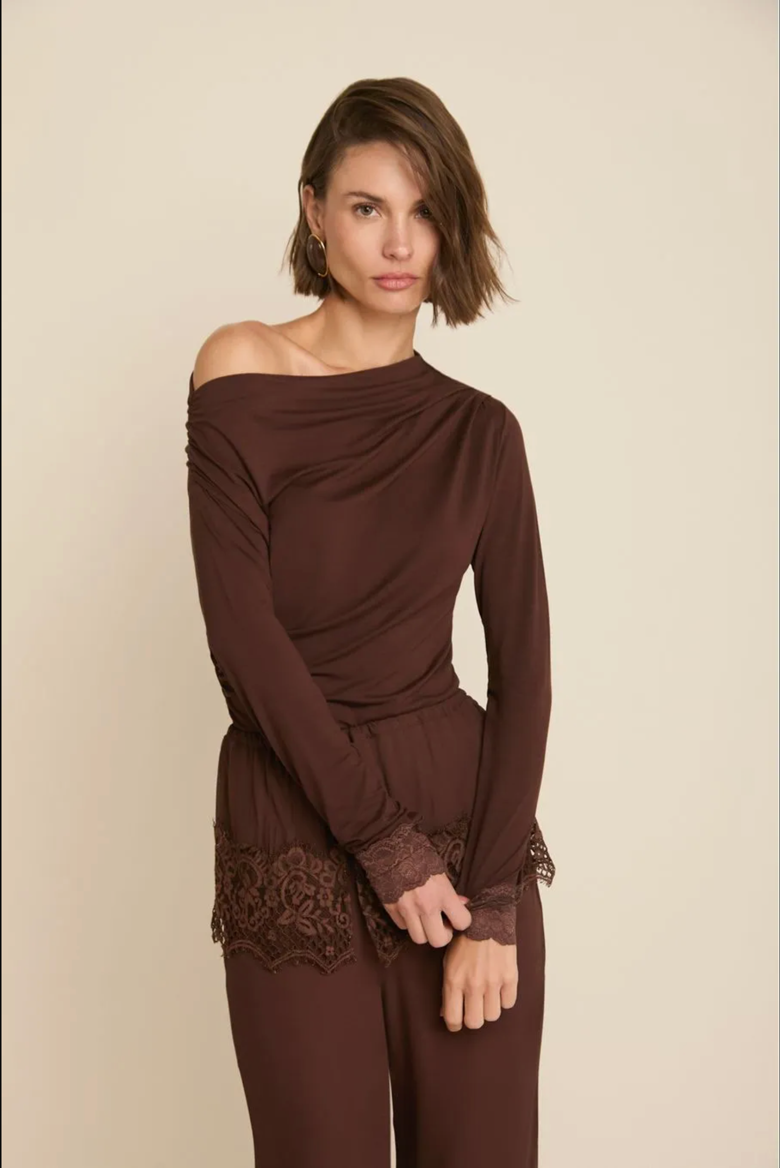 A woman modeling a dark brown off-the-shoulder long-sleeve top with matching brown pants, featuring lace detailing at the cuffs and hem, set against a plain beige studio background.