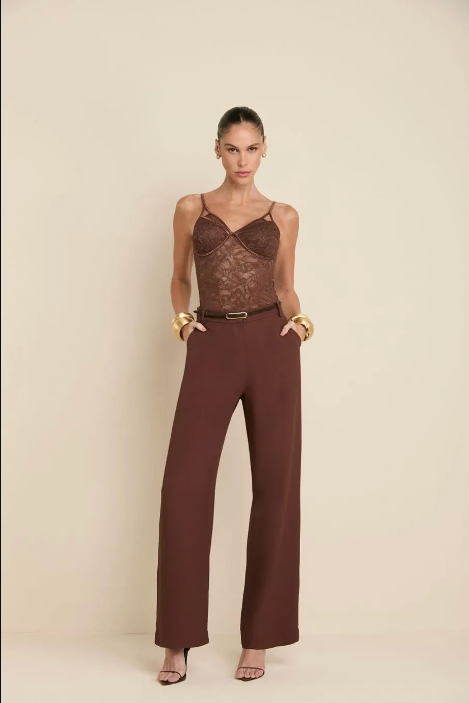 A model stands against a warm beige studio background wearing a brown lace camisole top, matching brown wide-leg pants, and gold bangles.