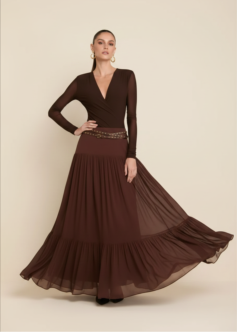 A woman modeling a long-sleeve maxi dress in dark brown, featuring a structured bodice, a decorative beaded waistband, and a flowing tiered skirt, set against a plain beige studio background.