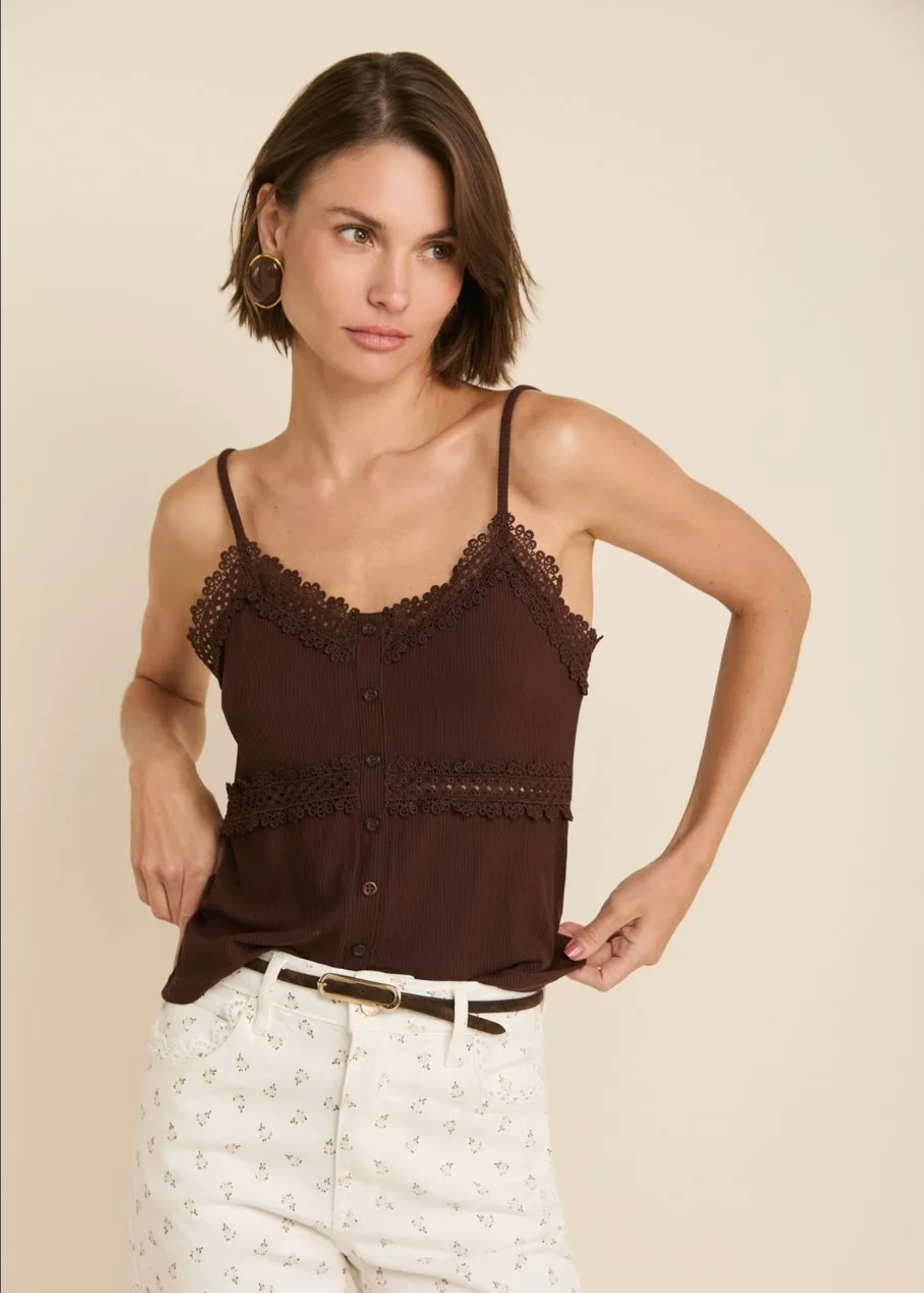 A woman modeling a dark brown camisole top with lace trim and button details, paired with white patterned pants, against a solid beige background.