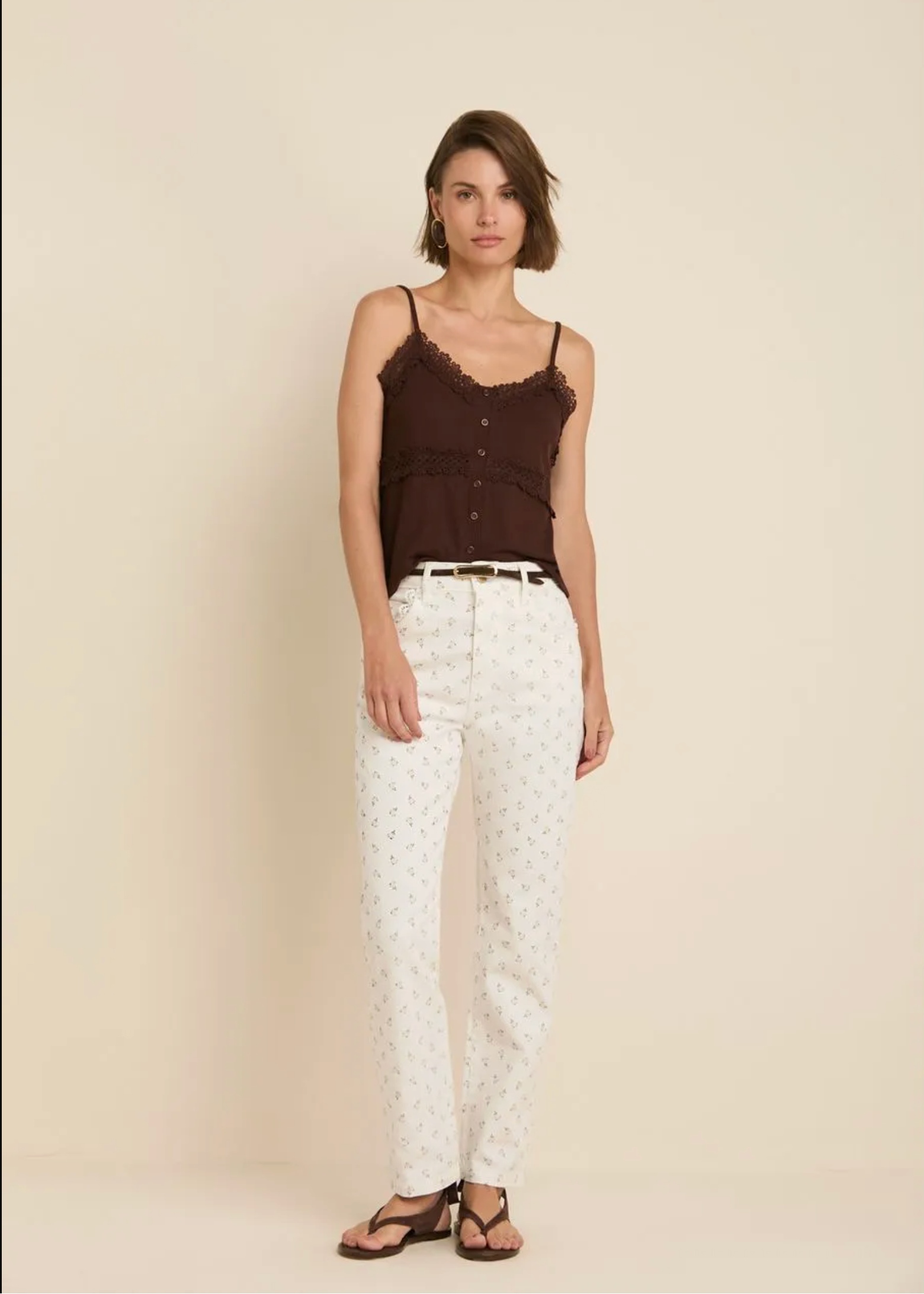 A woman modeling a dark brown ruffled camisole top and white pants with a small repeating pattern, standing against a plain beige studio background.
