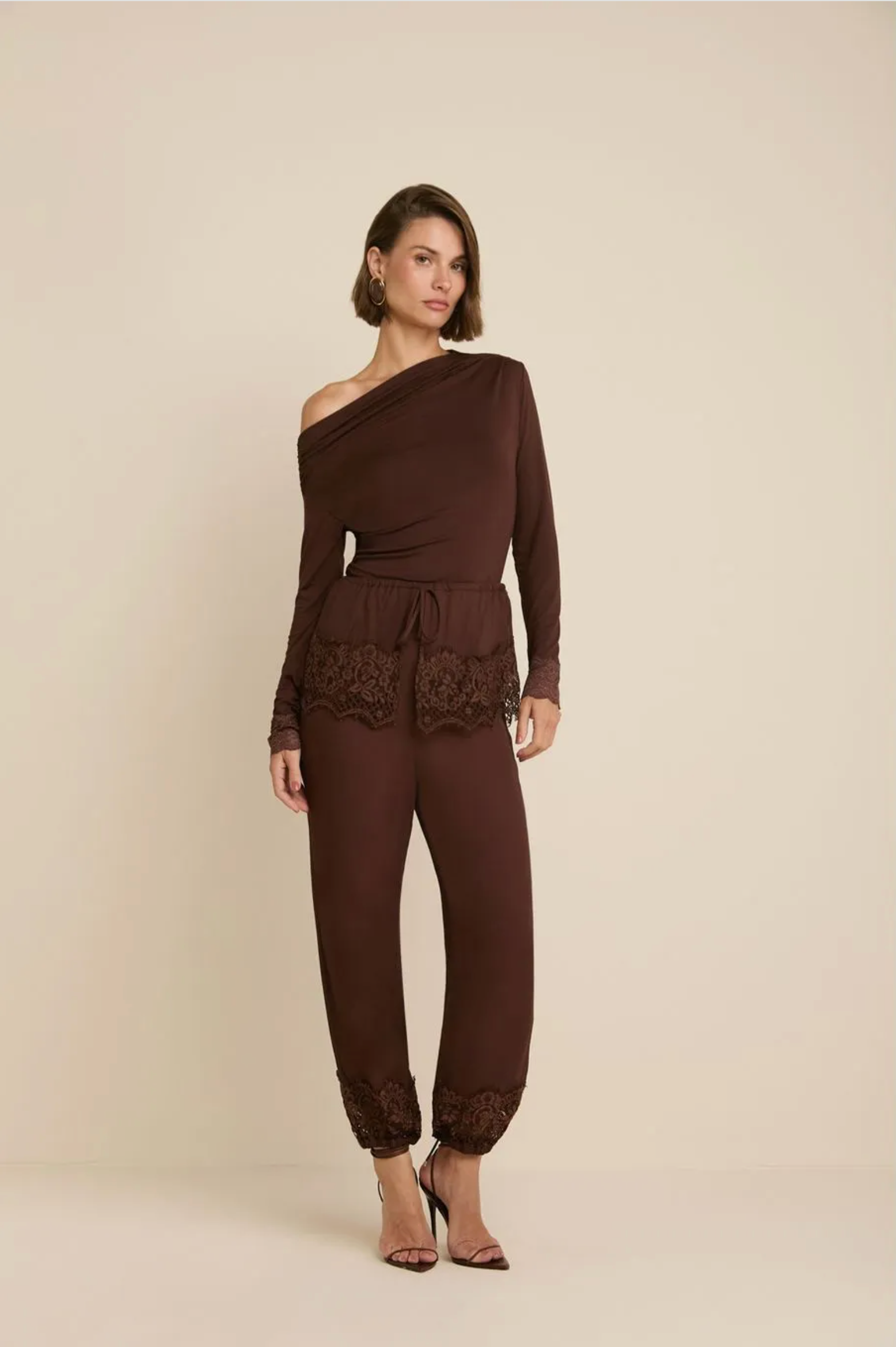 A woman models a dark brown one-shoulder jumpsuit with long sleeves, a tie waist, and lace trim details, paired with brown heeled sandals against a solid beige studio background.