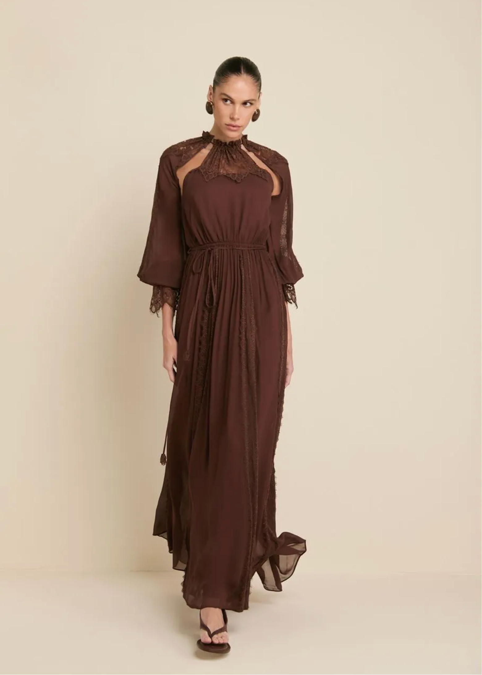 A woman modeling a long, dark brown midi dress with sheer lace details on the shoulders and sleeves, standing against a solid beige studio background.