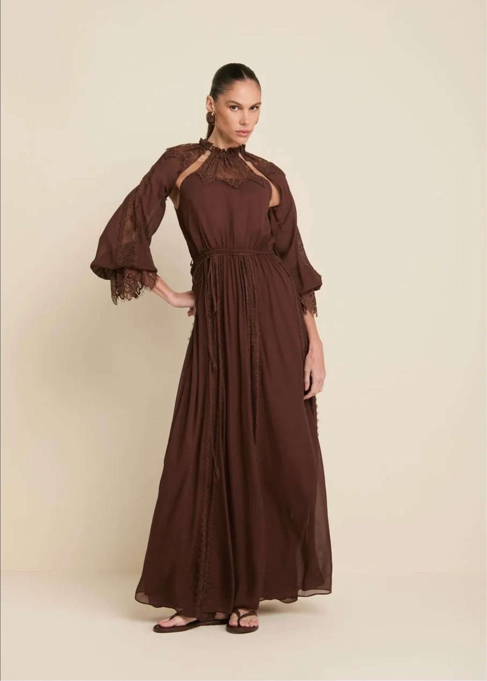 A woman modeling a long, flowing brown dress with sheer lace trim at the neckline and flared sleeves, standing against a plain beige studio background.