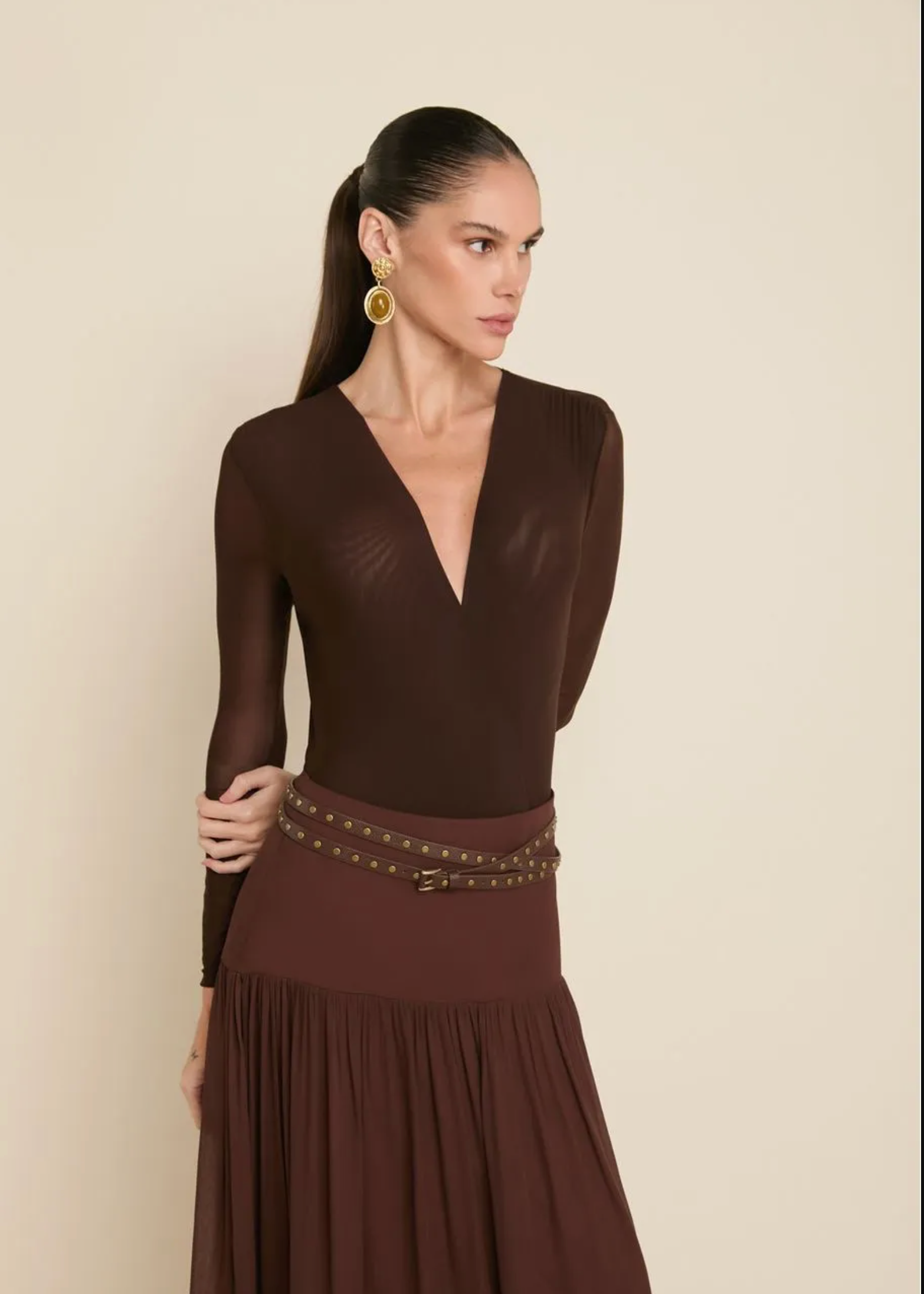 A woman modeling a dark brown long-sleeve dress with a deep V-neckline, sheer upper bodice, and a pleated skirt, accessorized with a studded belt. She is wearing large gold hoop earrings against a plain beige background.