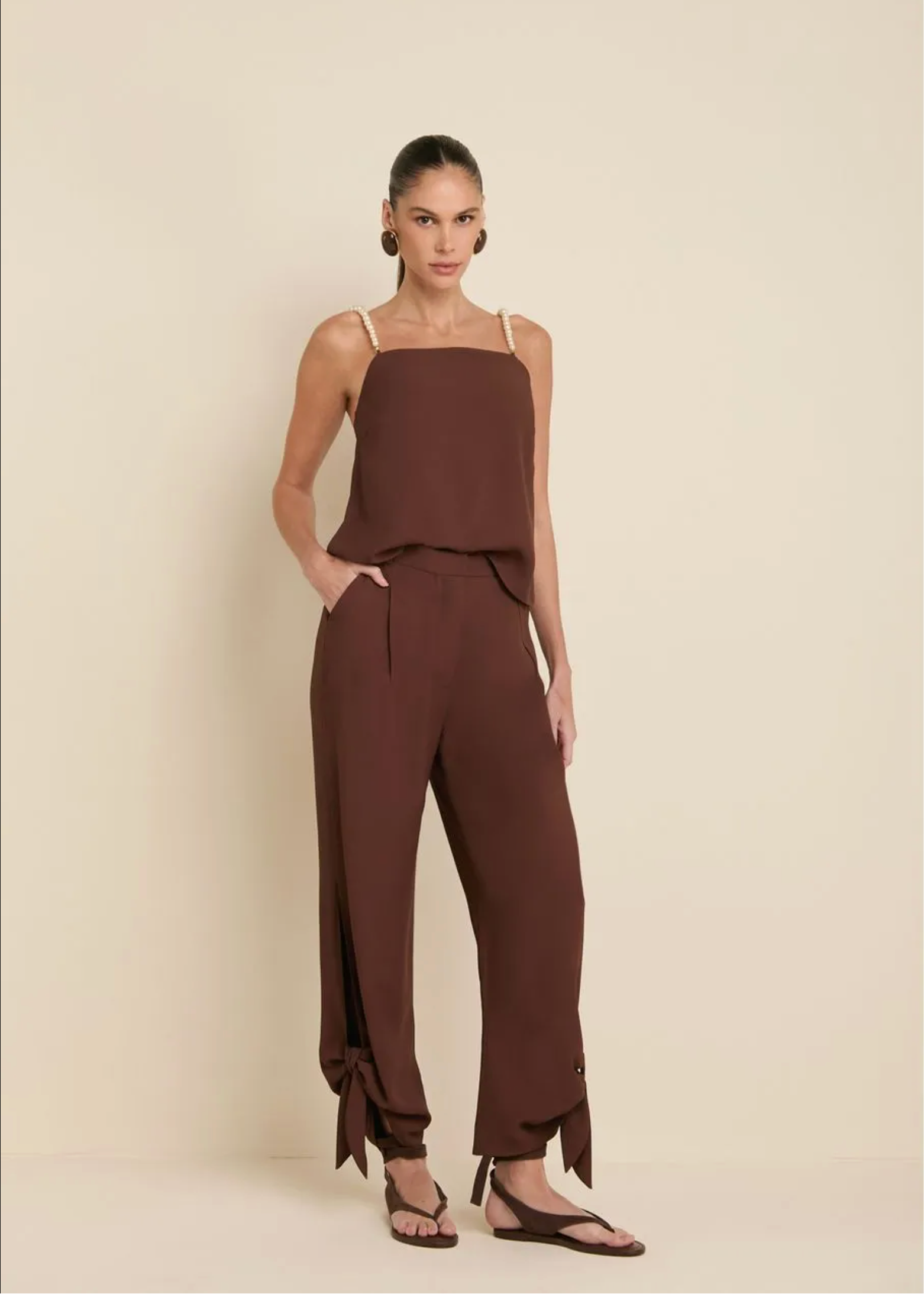A model wearing a matching two-piece brown set consisting of a sleeveless camisole top and wide-leg trousers, standing against a solid beige studio background.