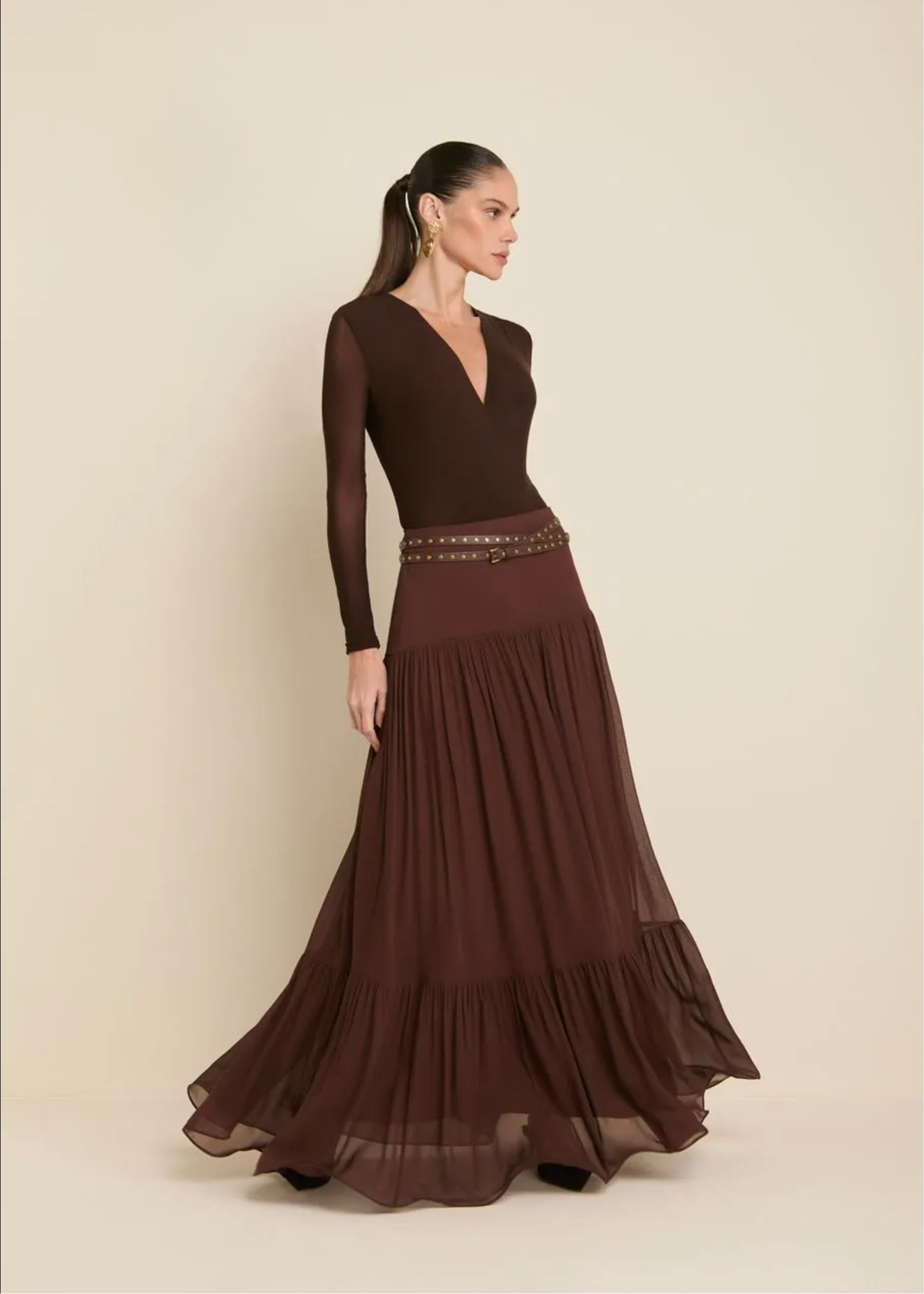 A woman modeling a long-sleeve maxi dress in dark brown, featuring a structured bodice, a decorative beaded waistband, and a flowing tiered skirt, set against a plain beige studio background.