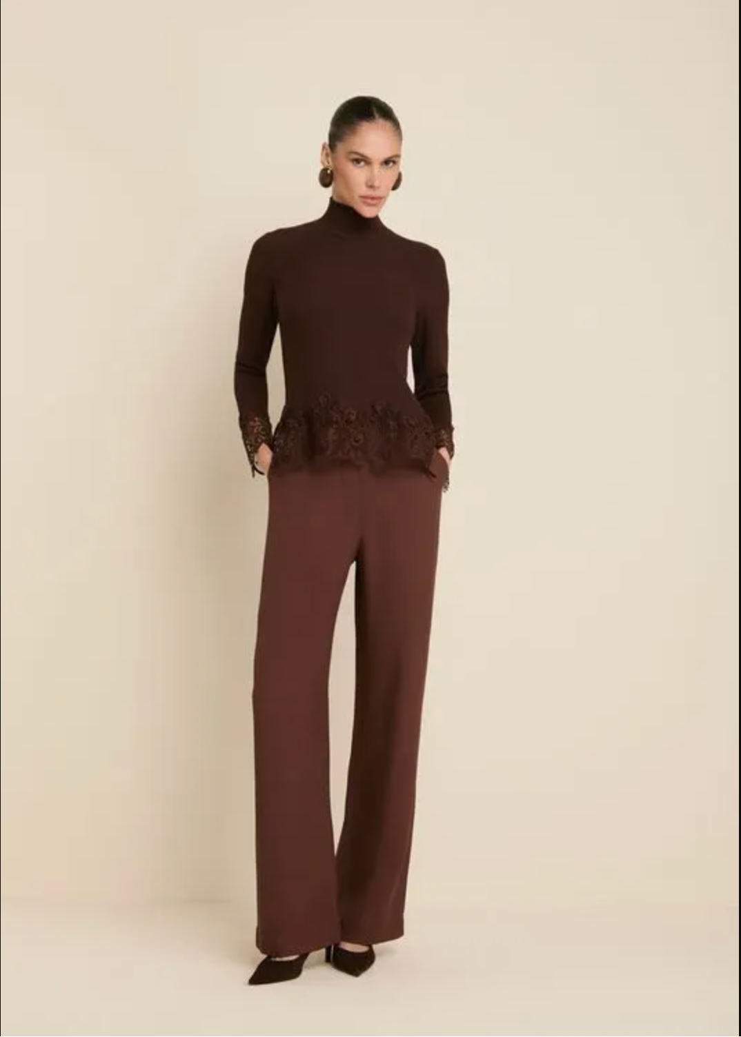 A model wearing a dark brown two-piece set consisting of a mock-neck long-sleeve top with a lace peplum hem and matching wide-leg trousers, standing against a solid beige background.