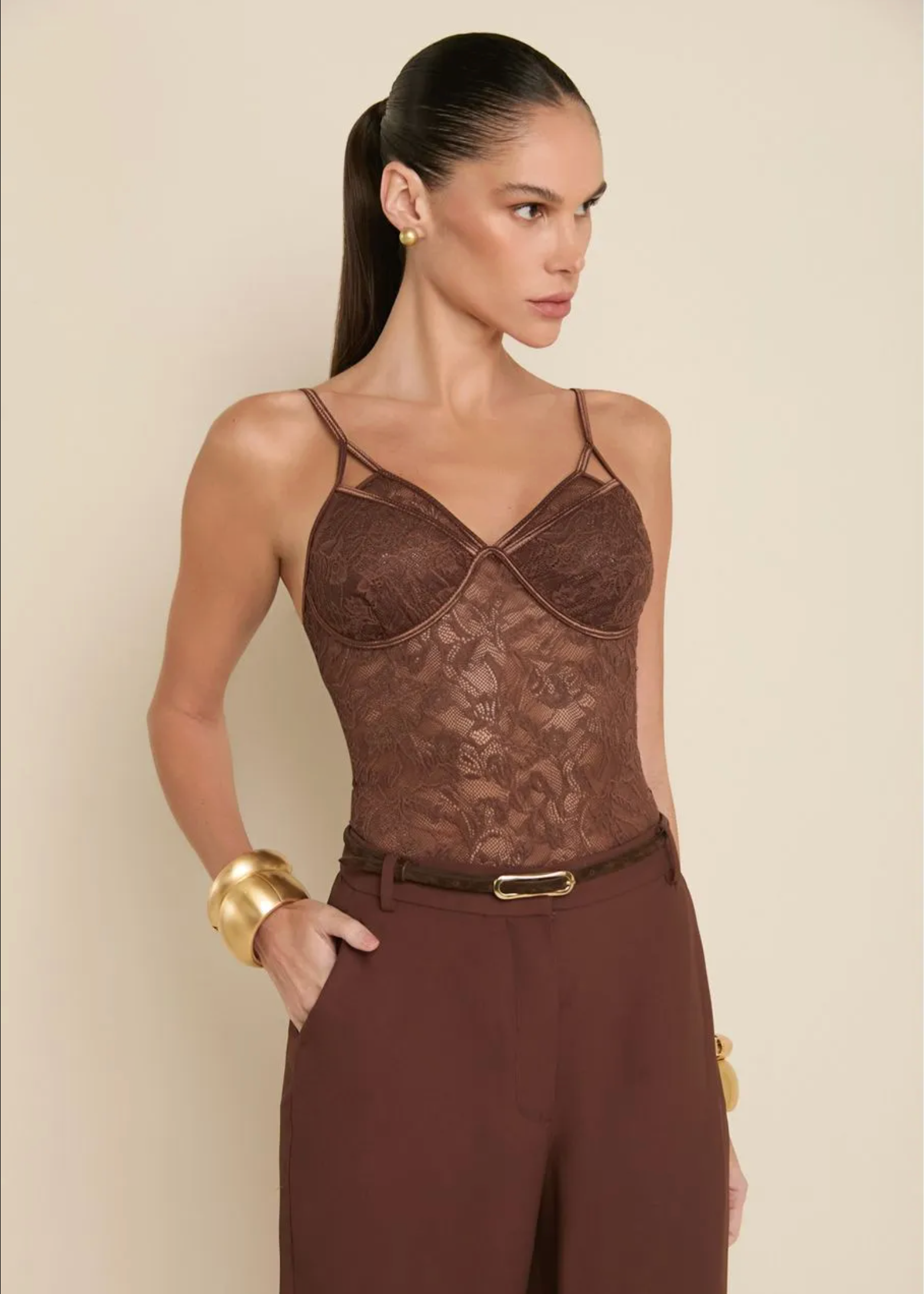 A woman models a brown lace camisole top with matching brown trousers, a thin brown belt, and gold jewelry against a plain beige background.