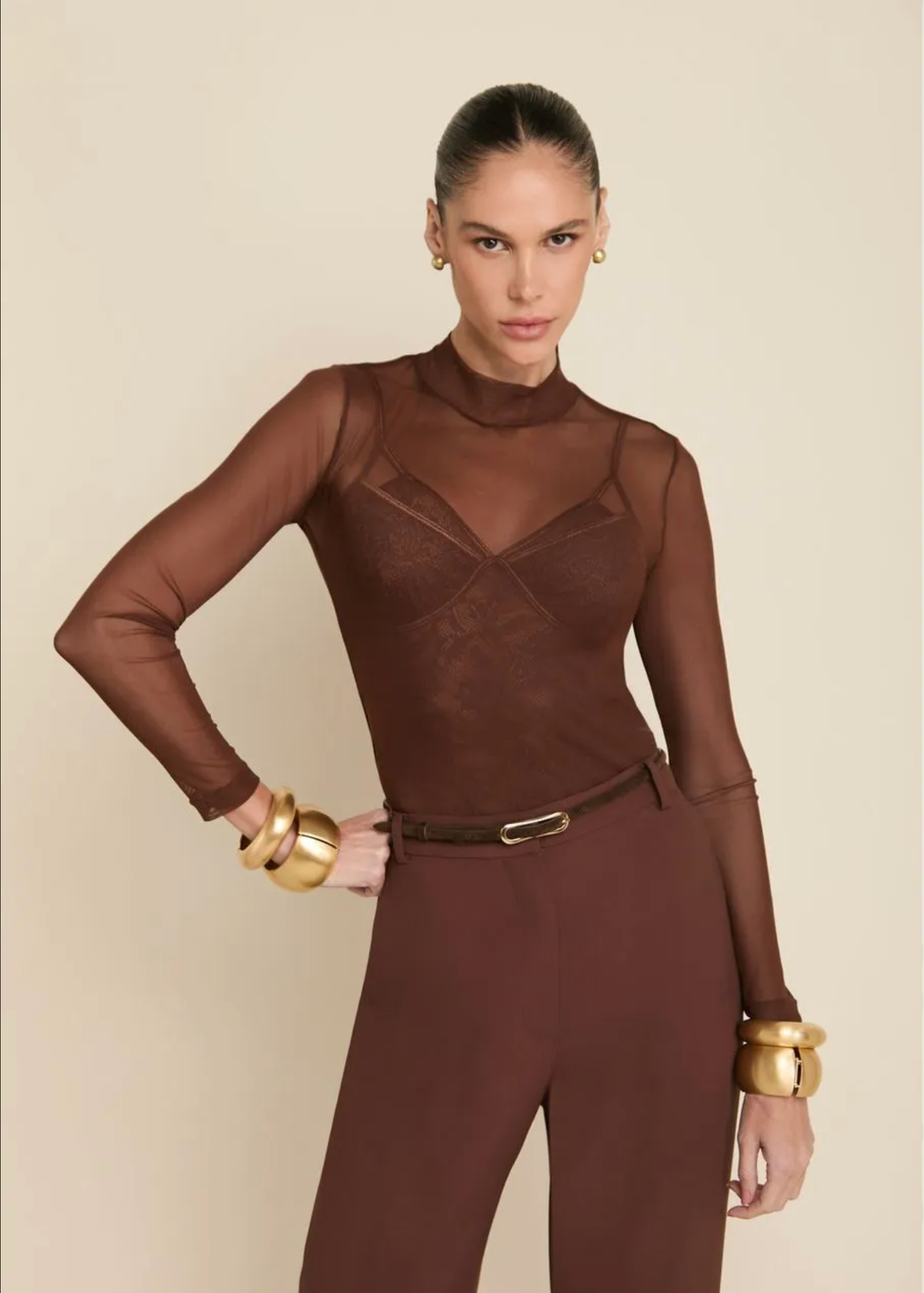 A model wearing a brown sheer mesh long-sleeve bodysuit with matching brown pants, styled with gold bracelets and a thin brown belt, against a solid beige background.