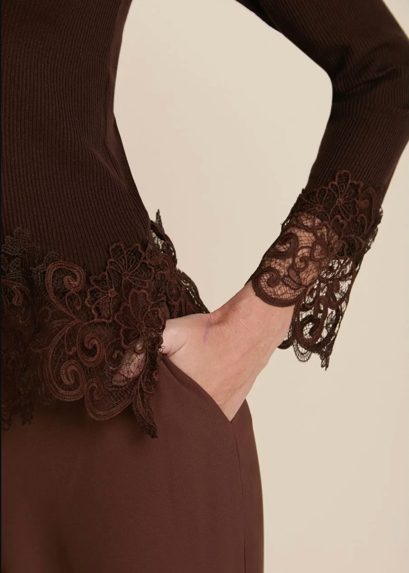 A woman modeling a dark brown mock-neck ribbed knit top with lace trim at the hem and cuffs, paired with matching brown trousers, against a solid beige background.