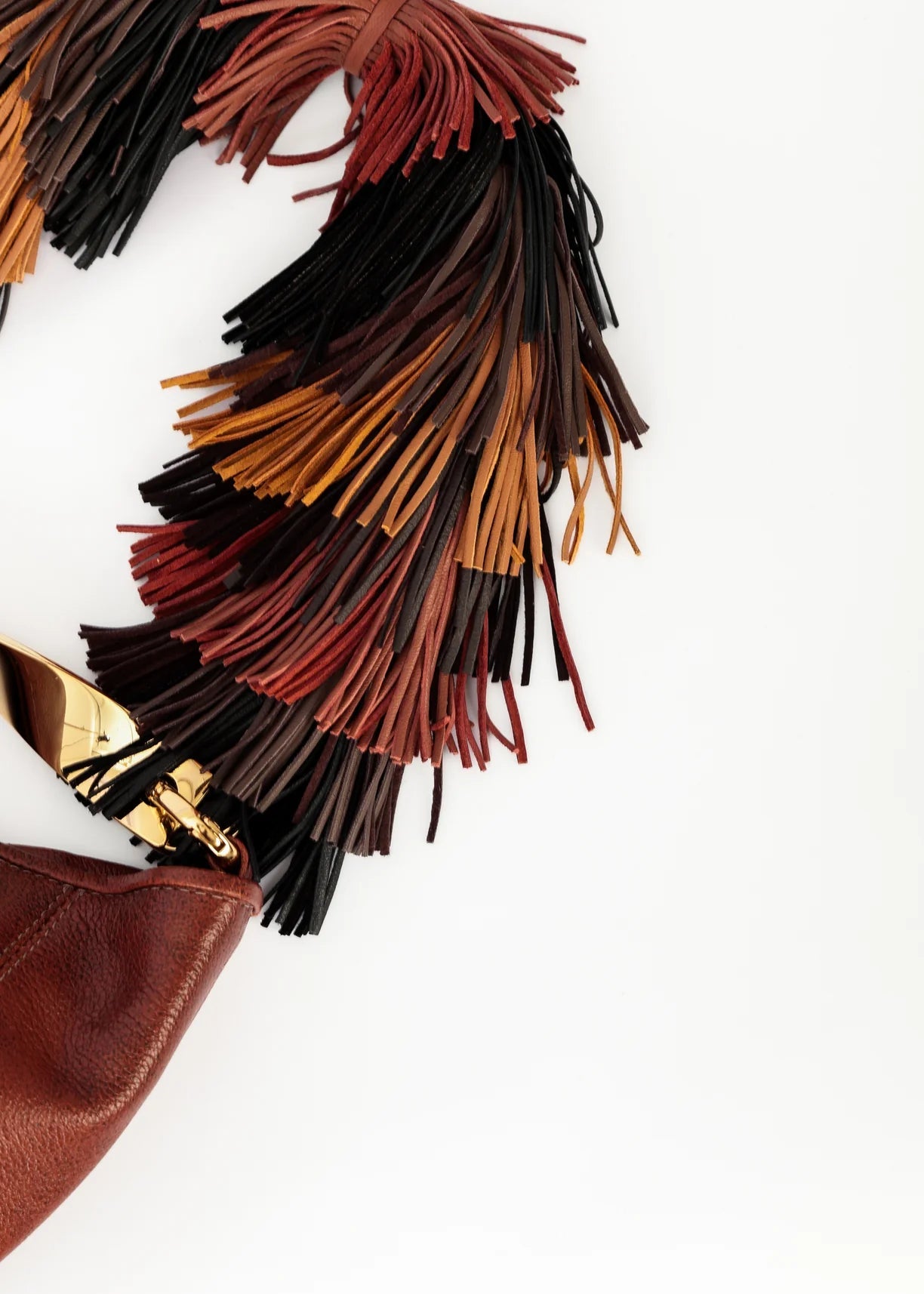 A close-up of a brown leather handbag corner featuring a dramatic, wide fringe tassel in layered shades of black, dark red, and tan, with gold-tone hardware, set against a plain white background.