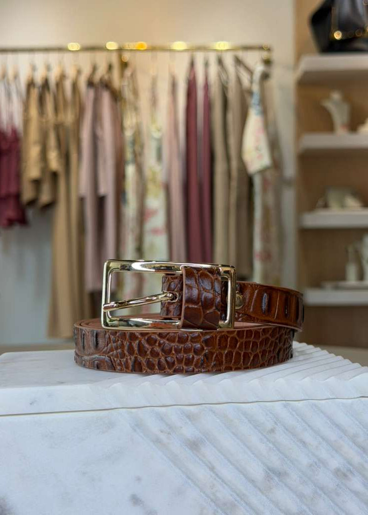 Brown crocodile-patterned belt with gold buckle on a marble surface, with clothing racks in the background.