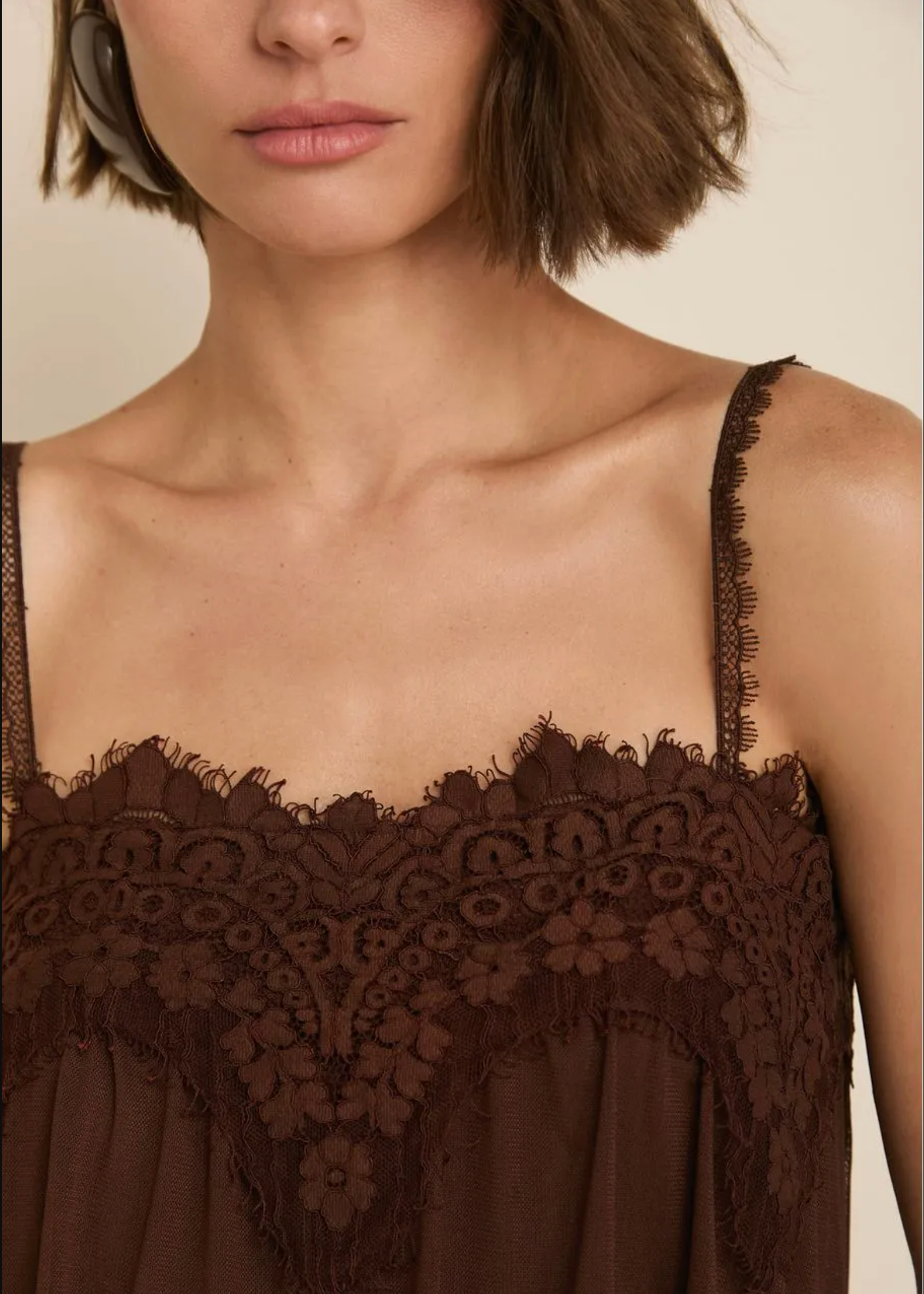 A close-up of a model wearing a dark brown camisole top with delicate floral lace trim and thin spaghetti straps against a warm beige background.