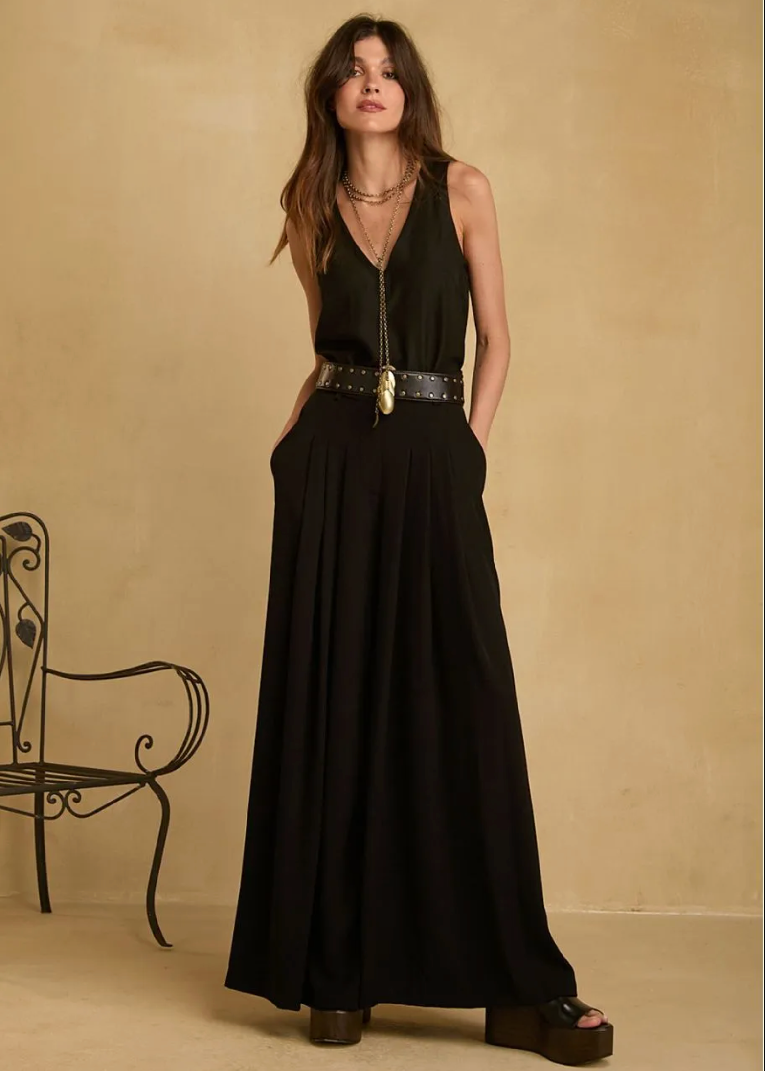 A woman models a black sleeveless maxi dress with a deep V-neckline, side pockets, and a wide black belt with gold studs and a gold pendant, set against a warm beige textured wall with a black metal chair on the left.
