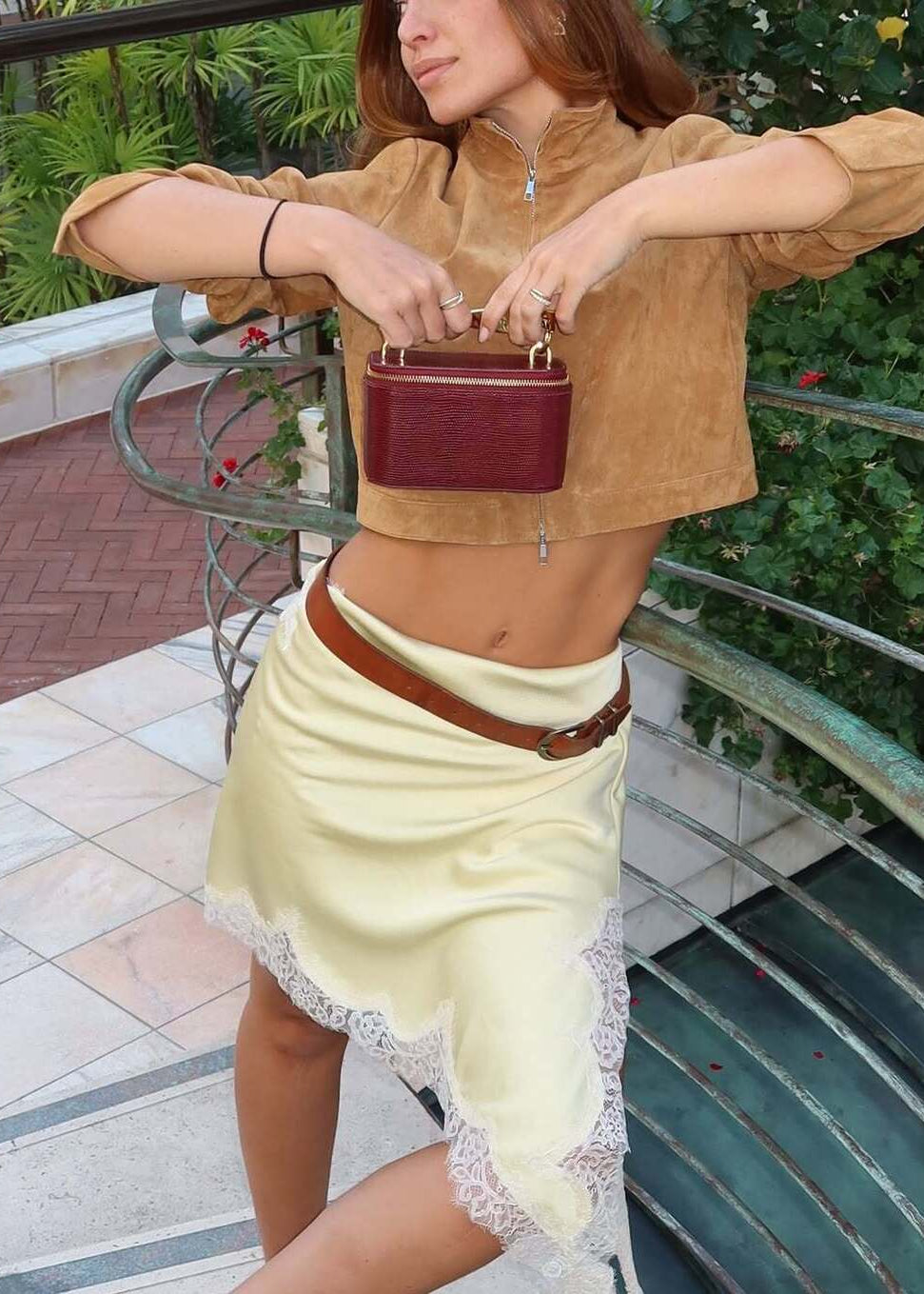 Woman holding a maroon handbag on a balcony with greenery in the background