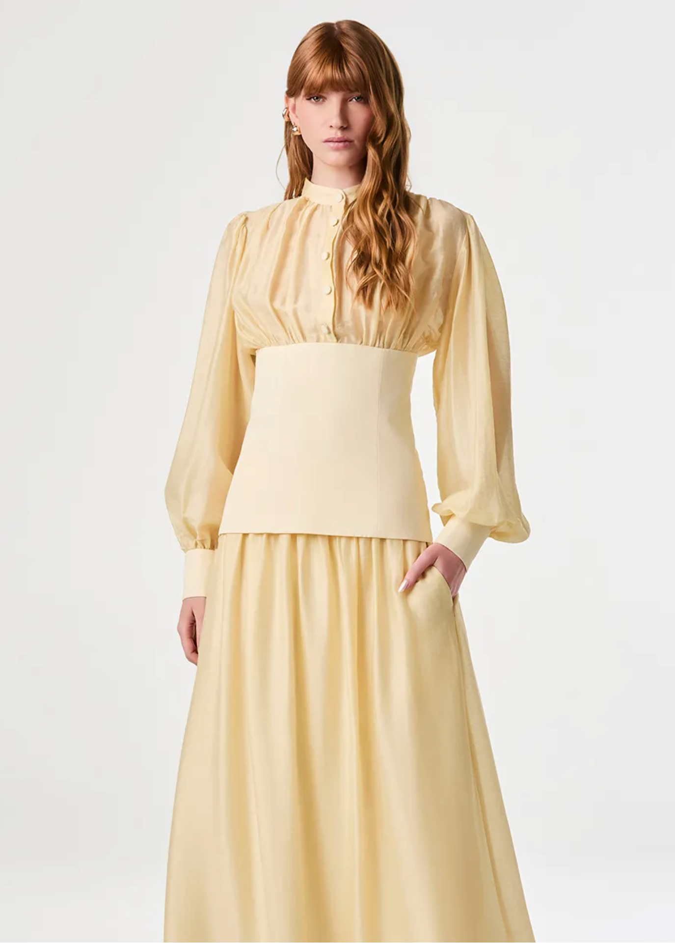 A model wearing a pale yellow two-piece dress with a long-sleeved blouse and a matching pleated skirt, photographed against a seamless light gray studio background.