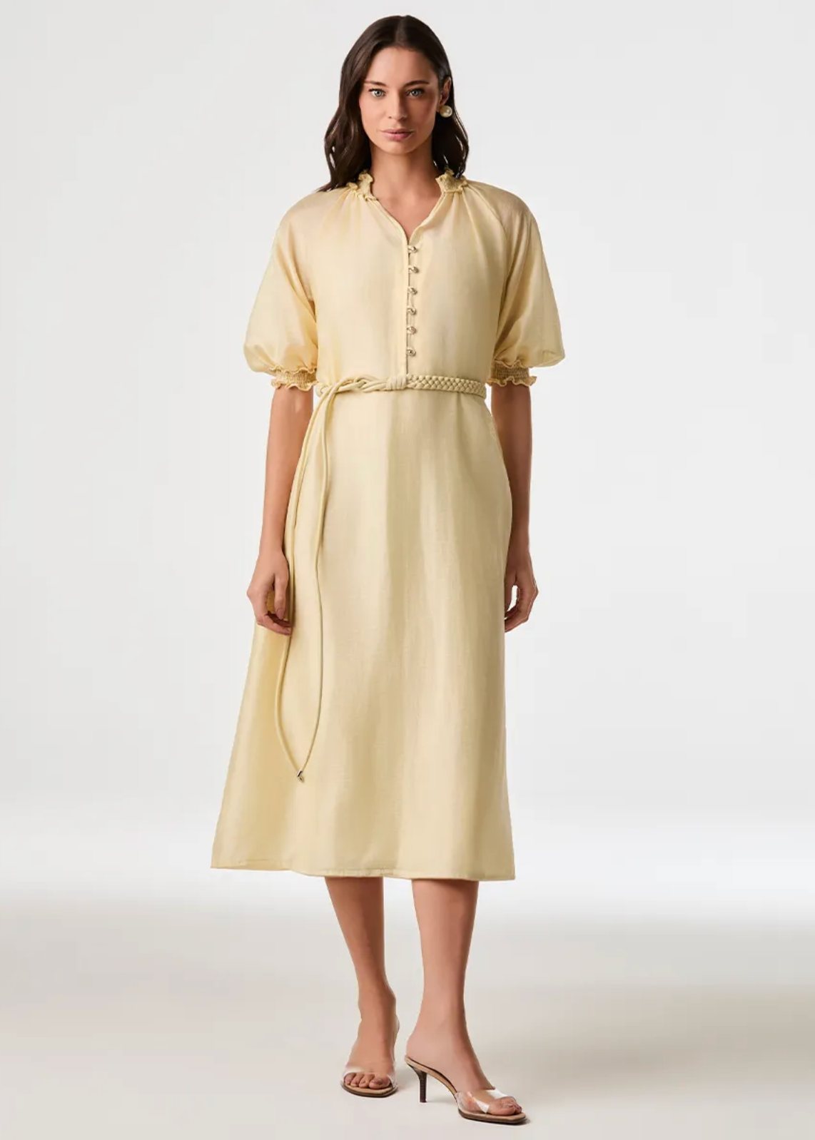A woman modeling a pale yellow midi dress with short puff sleeves, a buttoned neckline, and a drawstring waist, photographed against a clean light gray studio background.